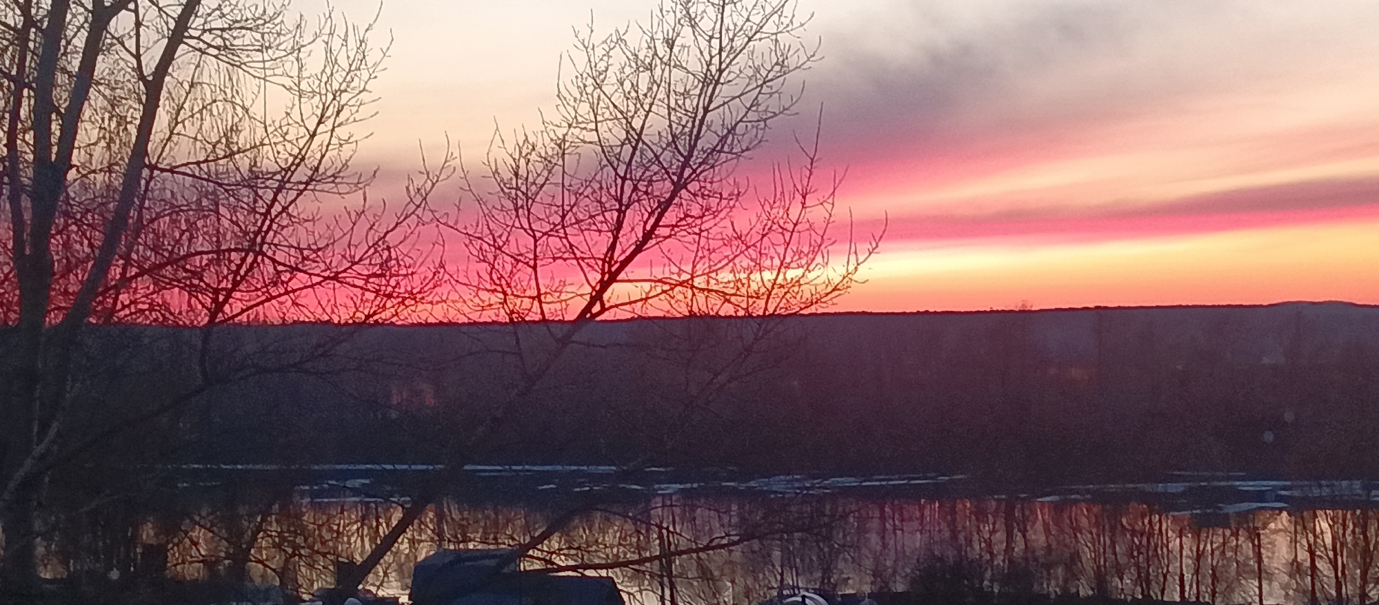 The image depicts a landscape scene at sunrise. Silhouetted, leafless trees are visible in the foreground. In the background, a vibrant sky with hues of pink, red, orange, and yellow, gradually fading to white, is seen above a dark, distant horizon. A body of water, possibly a river or lake, is reflecting the colors of the sky. The reflection also reveals the shadowy outlines of the trees.