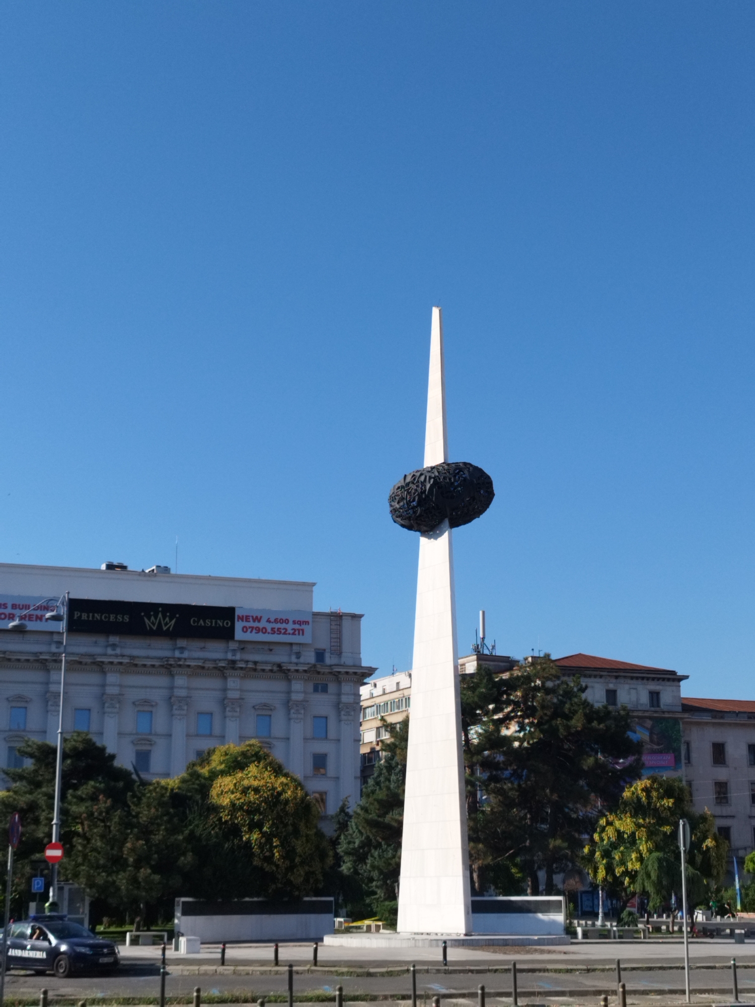 The image captures the Monumentul Renașterii, also known as the Spălătorul de Suflete (Soul Washer), located in Piața Revoluției (Revolution Square) in Bucharest, Romania. The monument is a tall, white, obelisk-like structure topped with a dark, irregular spheroid shape. In the background, a large, ornate building with classical architectural features stands prominently. A banner on this building advertises "PRINCESS CASINO" and mentions "NEW 4,600 sqm" along with a phone number. To the left of the main building, another building is visible with signage indicating "IS BUILDING" and "FOR RENT." Trees with lush green and yellow foliage are situated in front of and between the buildings. A dark-colored police car, marked "JANDARMERIA," is parked on the left side of the street in the foreground. The scene is set against a clear, bright blue sky, suggesting a sunny day. The ground in the foreground appears to be a paved area, likely a street or plaza, with various bollards and a curb.