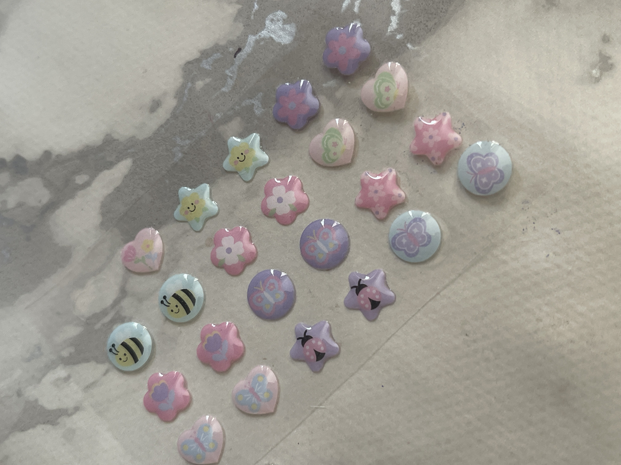 The image displays a collection of colorful, small, cartoonish stickers on a textured, light-colored surface. The stickers are arranged in a somewhat scattered pattern, with some overlapping. The shapes of the stickers include bees, flowers, hearts, butterflies, ladybugs, and stars. The colors are predominantly pastel shades of pink, purple, blue, and yellow. The surface appears to be a thin, possibly slightly wrinkled material with some darker, irregular smudges visible. The lighting is overhead and slightly diffused. There are no people or animals present in the image, and no discernible connection to the city of Tallinn, Estonia.