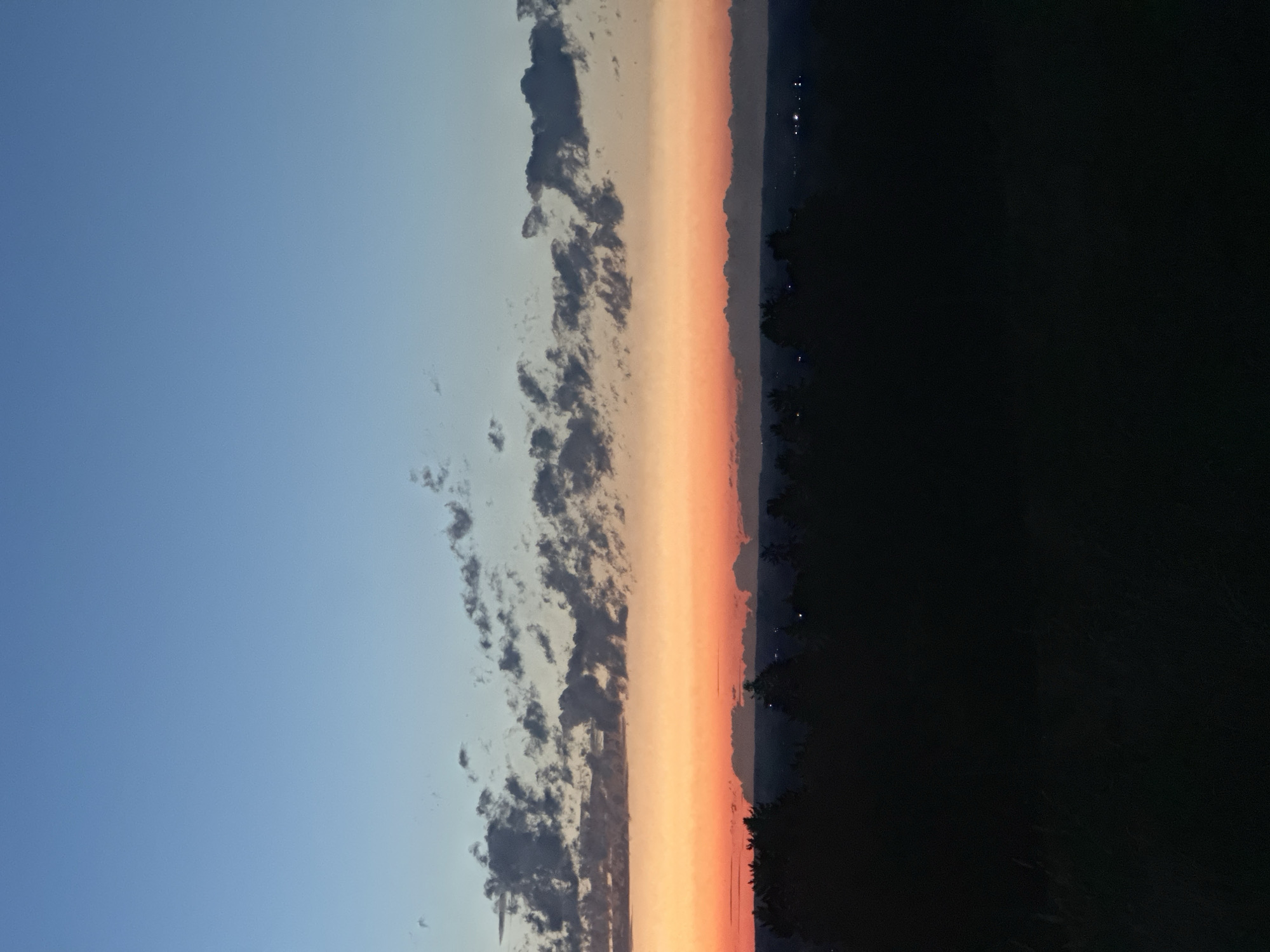 This is a photograph of a sunset over a forested area, likely taken from an elevated position. The sky transitions from a clear blue at the top to a band of orange and pink near the horizon, with scattered, dark clouds creating texture in the middle sky. Silhouetted against the colorful sunset is a dark band of trees, representing a forest or treeline. Beyond the trees, faint lights suggest a settlement or city in the distance. The foreground is dark and appears to be a grassy or vegetated area, also in silhouette. The overall impression is one of a natural landscape at twilight, with hints of human presence in the distance. The location in Amerang, Germany is not explicitly depicted within the image itself, but can be inferred from the prompt.
