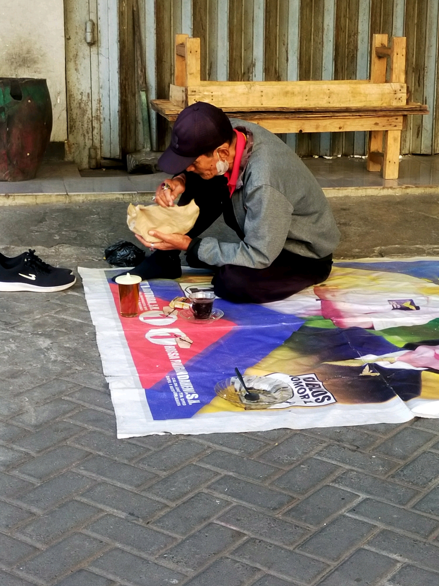 A person wearing a dark blue cap, a grey long-sleeved shirt, and a pink face mask is sitting on a colorful tarp on a brick-paved surface. They are holding a piece of food wrapped in brown paper. To the left of the person is a black athletic shoe. On the tarp, there is a glass of amber liquid, a teacup with dark liquid, and a small plate with a spoon and some remnants of food. In the background, there is a wooden bench and a weathered wooden door with vertical planks. The scene appears to be outdoors, possibly in a public area or marketplace in Surabaya, Indonesia, given the architectural style and the type of seating visible. The lighting suggests it is daytime.
