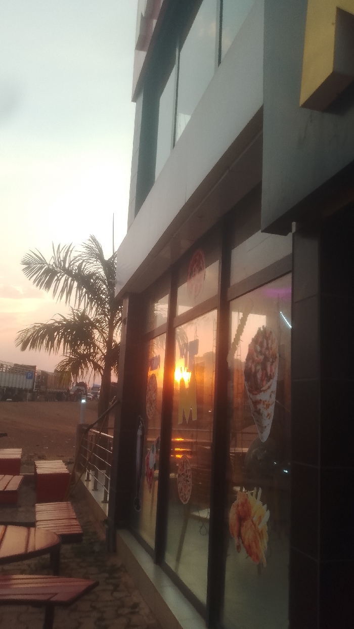 A building with large windows displays food advertisements. The setting sun creates reflections on the glass, blurring the images. Outside, wooden tables and a palm tree add to the ambiance. Across a sandy area, large vehicles are visible in the distance under a bright sky.
