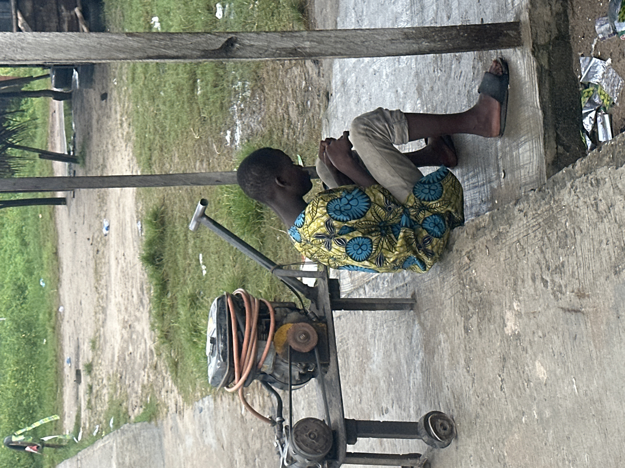 In Badagry, Nigeria, a young Black male, wearing a brightly colored yellow and blue patterned shirt and light-colored trousers, is seated on a concrete curb. He is looking down at his hands, which are clasped together. Beside him is a piece of equipment that appears to be a portable generator with a coiled orange hose. The ground is a mixture of concrete and dirt, with patches of green grass and scattered litter. In the background, there are wooden structures and palm trees, suggesting a semi-rural or informal settlement environment. The scene is outdoors, with natural lighting.