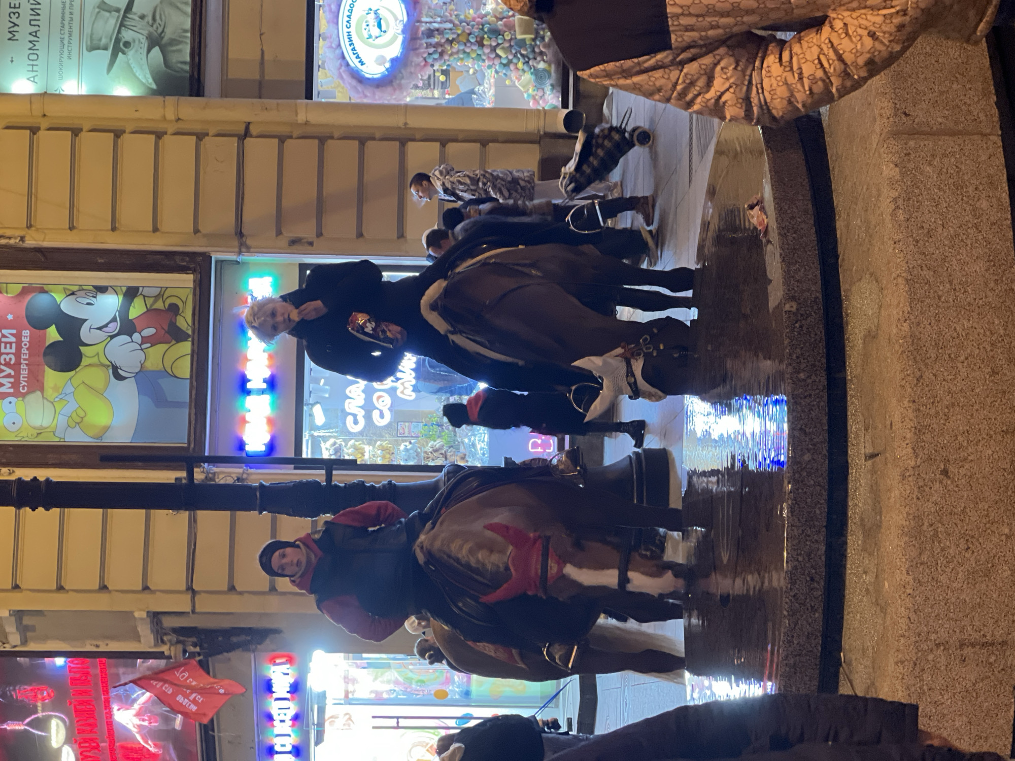 The image shows a scene on a street in Saint Petersburg, Russia, at night. Several people are riding horses on a wet, reflective pavement. There are shops and illuminated signs along the street, including one for a "Museum of Anomalies" and another for a "Museum of Superheroes" featuring Mickey Mouse. The street is lit by neon signs and streetlights, creating a bright, urban environment. The reflections on the wet pavement add to the dynamism of the scene.