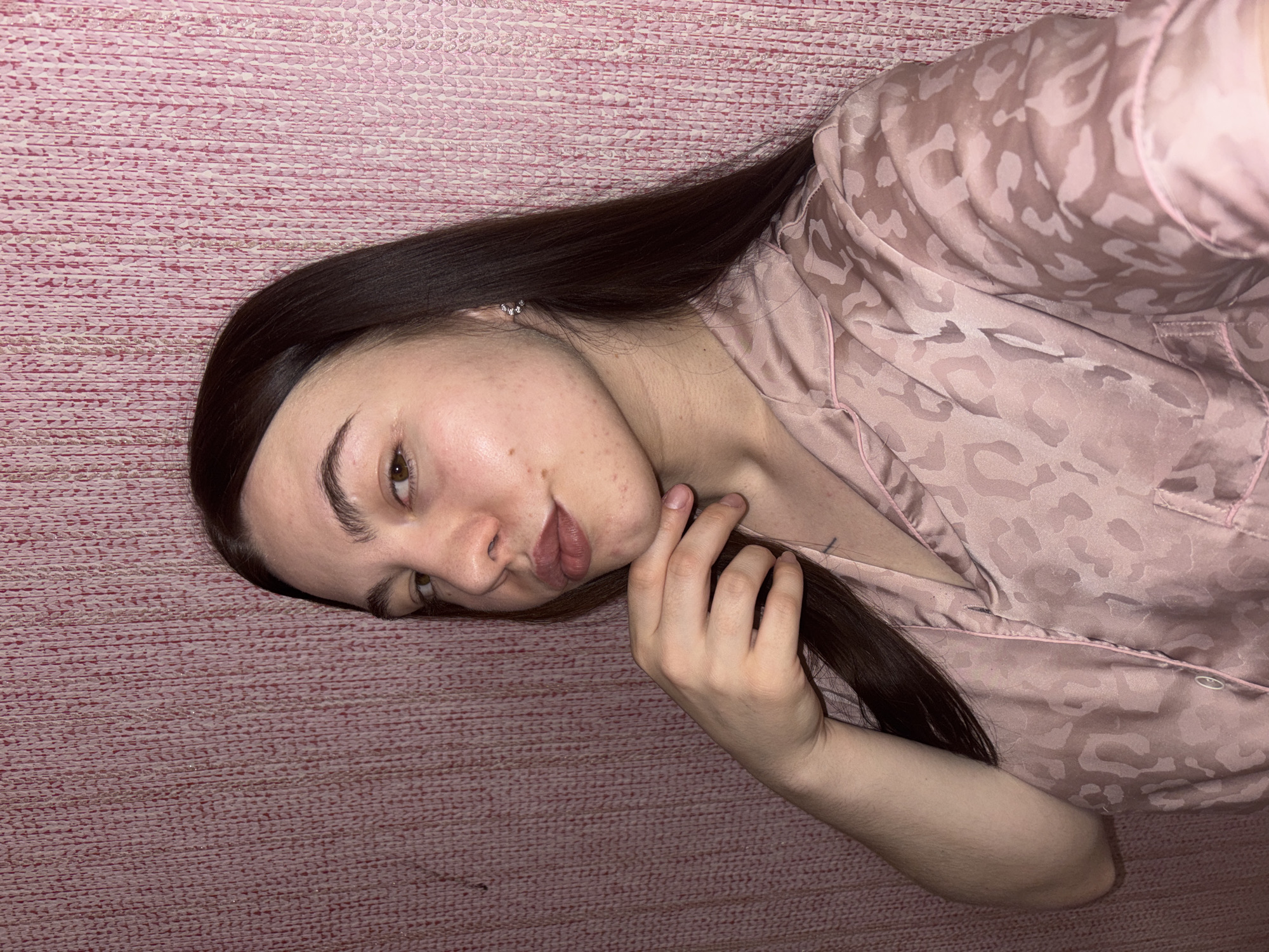 A young woman with long, dark brown hair and light skin is captured in a close-up, eye-level shot, positioned on her side with her head tilted. She has dark brown eyes and full lips, showing a slight pout or smirk. Her skin has some visible freckles or blemishes on her cheeks and forehead. She is wearing a light pink, satin-like pajama top with a subtle, lighter pink leopard print pattern. A small, sparkling earring is visible in her left ear. Her right arm is bent, and her hand is resting near her face, with unpolished nails visible. A faint dark marking, possibly a small tattoo, is visible on her upper left chest/shoulder area beneath her pajama top. The background is a textured pink surface, resembling wallpaper or a woven fabric, which she appears to be lying against or on. The lighting suggests an indoor setting. No specific time of day is discernible, and no visible text is present. The overall impression is relaxed and casual, likely a self-portrait taken in Perm, Russia.