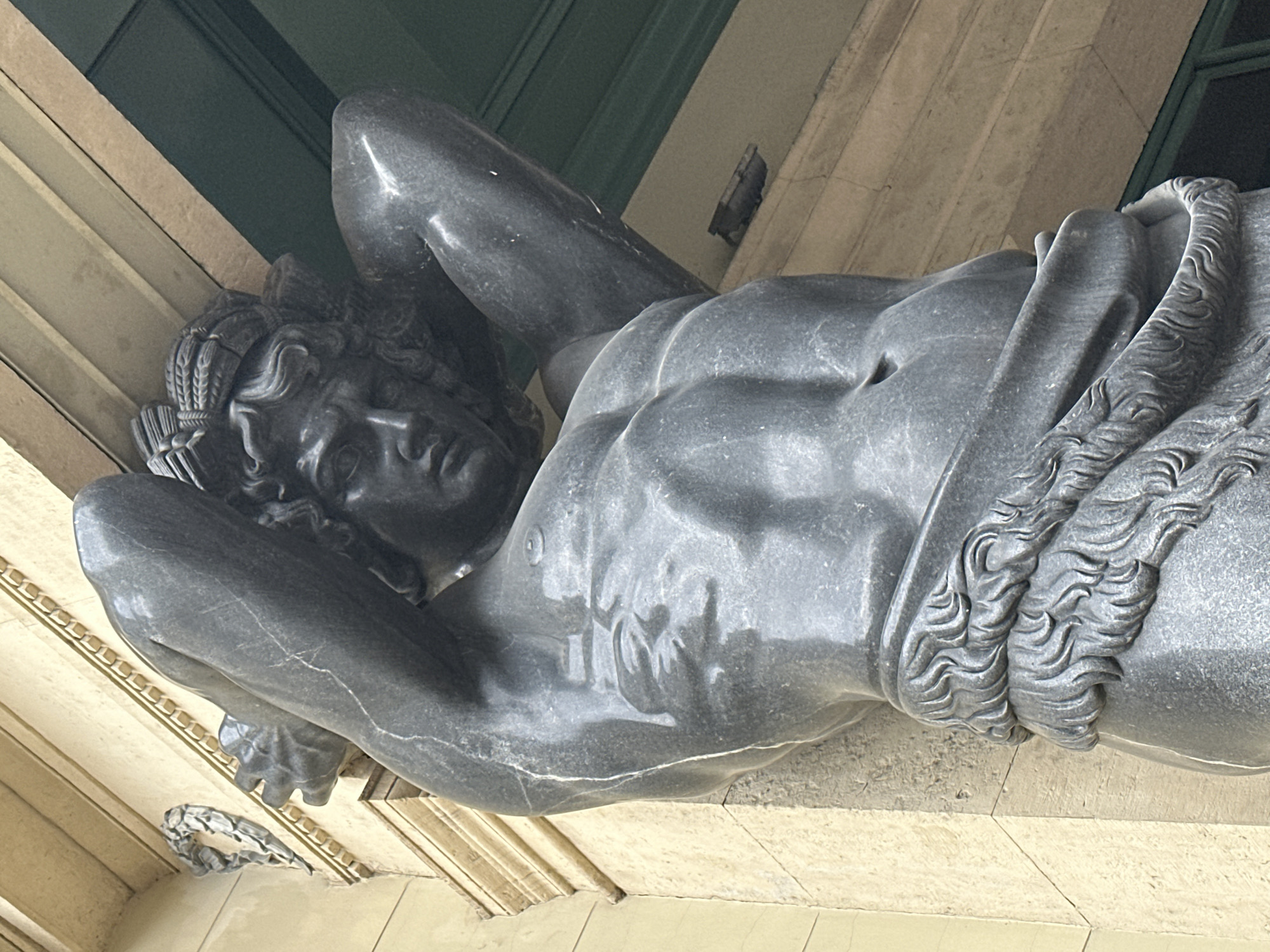 The image is a close-up, angled shot of a dark gray stone sculpture of a muscular male figure. The sculpture appears to be a statue of Atlas, as it is depicted in a position of carrying a great weight, with its arms raised above its head and its body contorted. The figure is detailed, with clearly defined muscles and an expressive face. It wears a laurel wreath on its head. The stone has a polished surface with some visible veins and cracks, indicating age or the natural properties of the material. The sculpture is positioned outdoors, as suggested by the muted natural lighting and the architectural elements visible in the background. These include a light-colored stone facade with decorative molding and a dark green architectural feature, possibly a window or a door, in the upper part of the frame. The scene is static, with no apparent activity taking place beyond the depiction of the figure's pose. The lighting suggests it could be daytime, possibly overcast or in shade, as there are no harsh shadows. The overall impression is one of strength, endurance, and classical artistry.