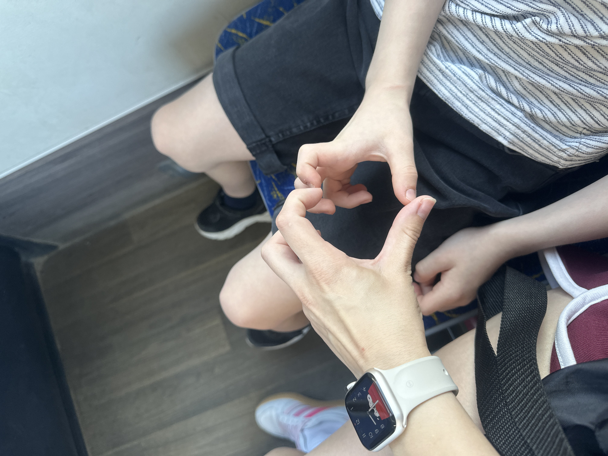 The image shows two people making a heart shape with their hands. One person is wearing a white Apple Watch, black shorts, and pink and white sneakers, while the other person is wearing a white and blue striped shirt and black shorts with black shoes. The location appears to be a bus or train, with blue seating visible. The exact location in Perm, Russia, is unknown without additional information, as only the interior of public transport is visible.