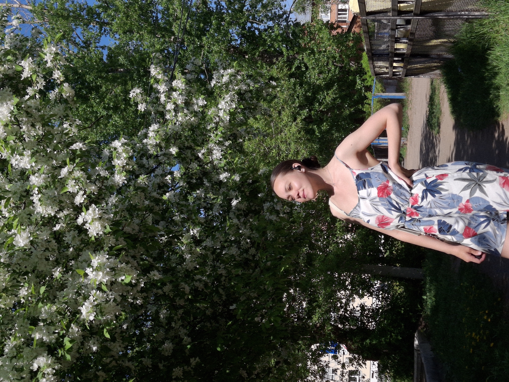 A woman stands in a park in Chaikovsky, Russia. She is wearing a light-colored dress with floral patterns. Behind her is a blooming tree with white flowers. In the background, there are buildings and a fence. The scene is bathed in sunlight. A dirt path is present with grass growing along side of it.