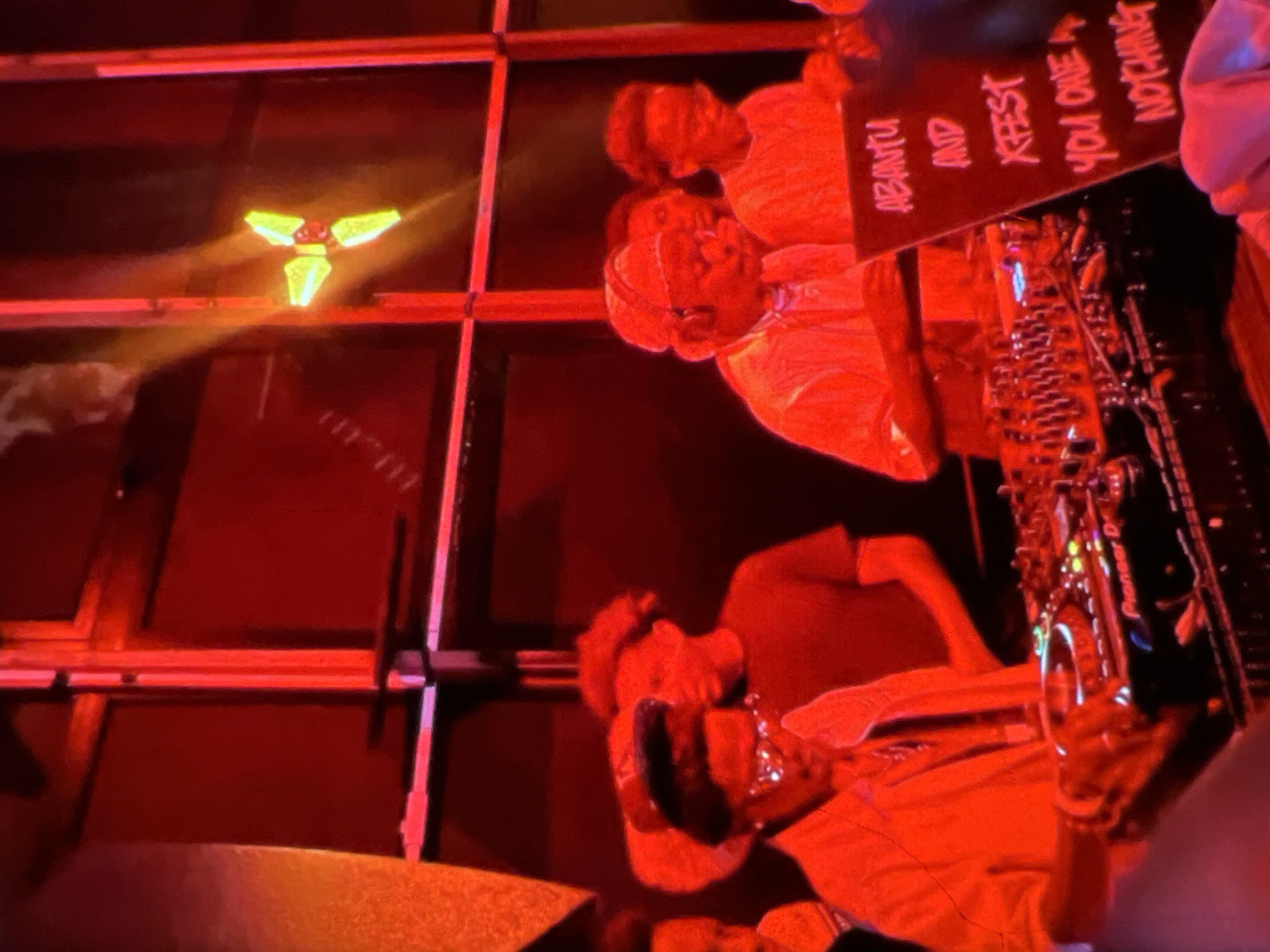 The image depicts a dimly lit indoor scene, likely a nightclub or event space, bathed in intense red lighting. In the foreground, a person wearing glasses and a hat is operating DJ equipment, with a Pioneer DJ mixer prominently visible. They appear to be in the process of mixing music, with one hand raised. To the right of the DJ, a sign with white lettering on a black background reads "ABANTU AND XFEST YOU OWE A NOTHING". Several other individuals are visible in the background, some wearing headphones and appearing to be part of the event's crew or audience. The overall atmosphere is energetic and suggestive of a live performance or party. The presence of "XFEST" suggests this event might be part of a festival or celebration. While the image does not provide explicit clues to the city, the text on the sign "ABANTU" hints at potential cultural or artistic themes that could be relevant to Lagos, Nigeria, given the prevalence of music festivals and creative events in the city.