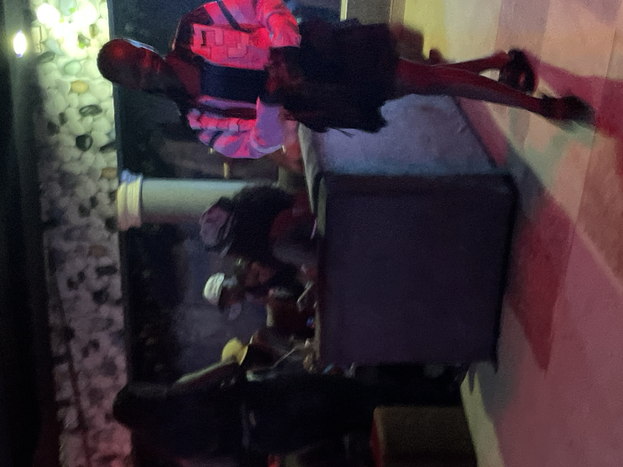 The image depicts an indoor scene at night, likely in a dimly lit venue in Durumi, Nigeria. The lighting is predominantly artificial, with red and pink hues casting shadows on the floor. In the foreground, a young woman is walking away from the camera, wearing a dark skirt, dark sandals, and a colorful jacket with pink and white accents. She is holding a dark bag in her right hand. Behind her, several other individuals are visible in the midground, seated at a low table or counter. One person in a white hat is partially visible. The background shows a textured wall with stones or rocks and some artificial lighting. To the left, a dark silhouette of another person stands. The overall atmosphere suggests a social gathering or an entertainment setting.