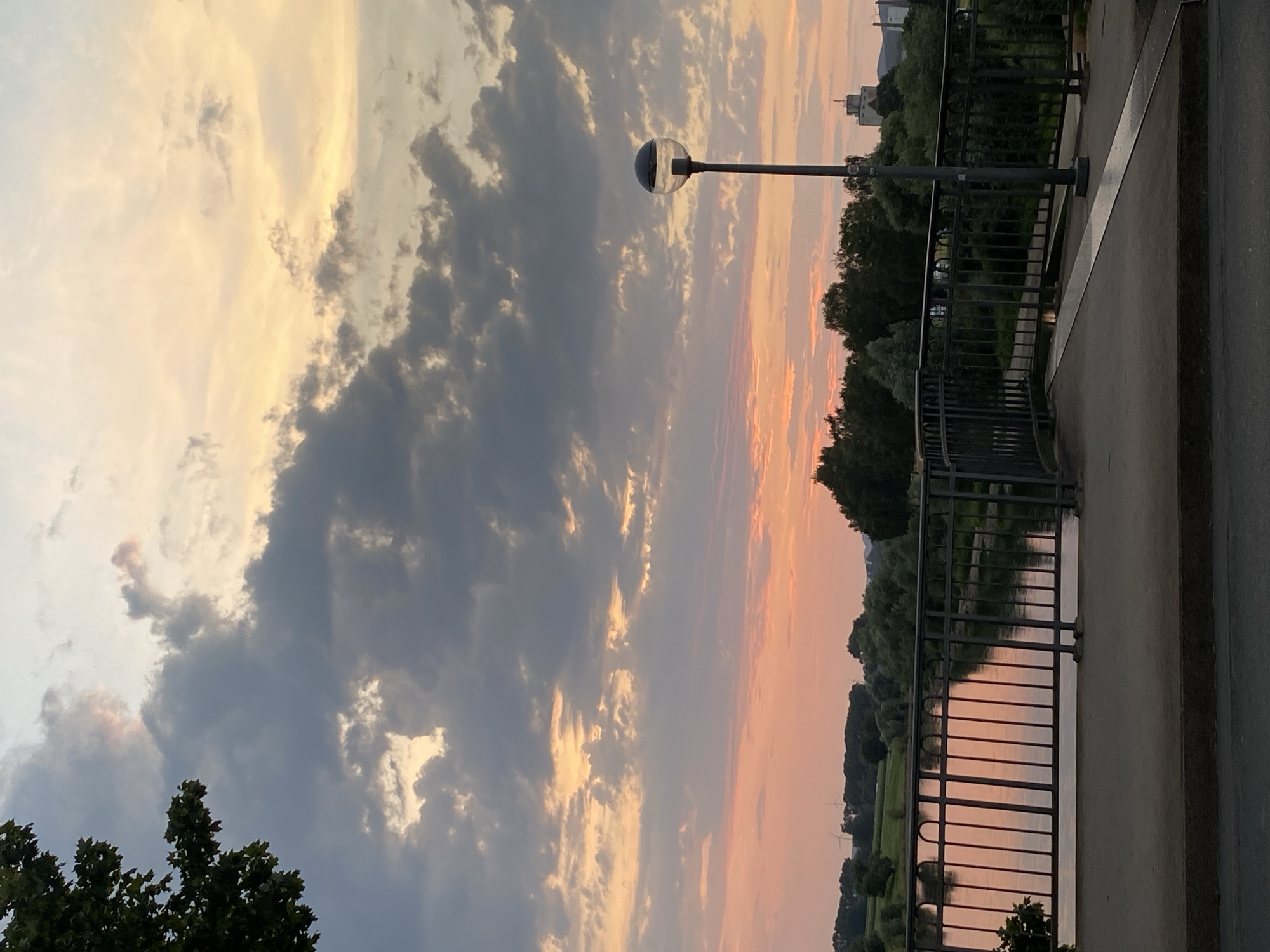 A wide-angle, vertical photograph captures a sunset over the Weser River in Rinteln, Germany. The sky is a dramatic mix of orange, pink, and grey clouds, with hints of yellow near the horizon. In the foreground, a metal railing and a walkway lead the eye towards the river. The river's surface reflects the colorful sky and the surrounding greenery. On the far bank, a line of trees separates the river from fields and distant buildings, including what appears to be an industrial facility and a church spire. A lone lamppost with a spherical head stands to the right of the frame. The overall atmosphere is serene and picturesque, highlighting the natural beauty of the area as day transitions into night.