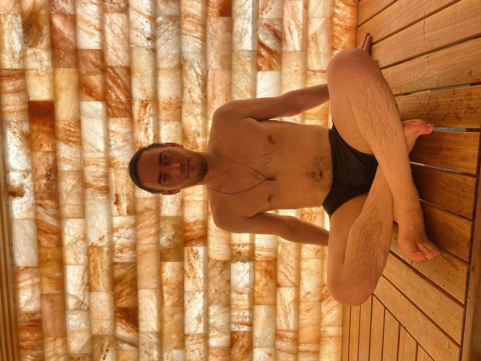 A middle-aged man is sitting on a wooden platform inside what appears to be a salt cave or sauna. The man is fair-skinned, has a beard and short dark hair, and is wearing dark-colored swim trunks or underwear. He is shirtless and has a thin necklace around his neck. His legs are crossed, and his right arm is extended, resting on the wooden planks. The walls of the room are made of rectangular blocks of salt, which are backlit and appear to be various shades of white, orange, and brown. The wooden platform is constructed of parallel planks and is also illuminated. The scene appears to be indoors in a spa or wellness facility, although the specific location in Votkinsk, Russia, is not discernible from the image itself.