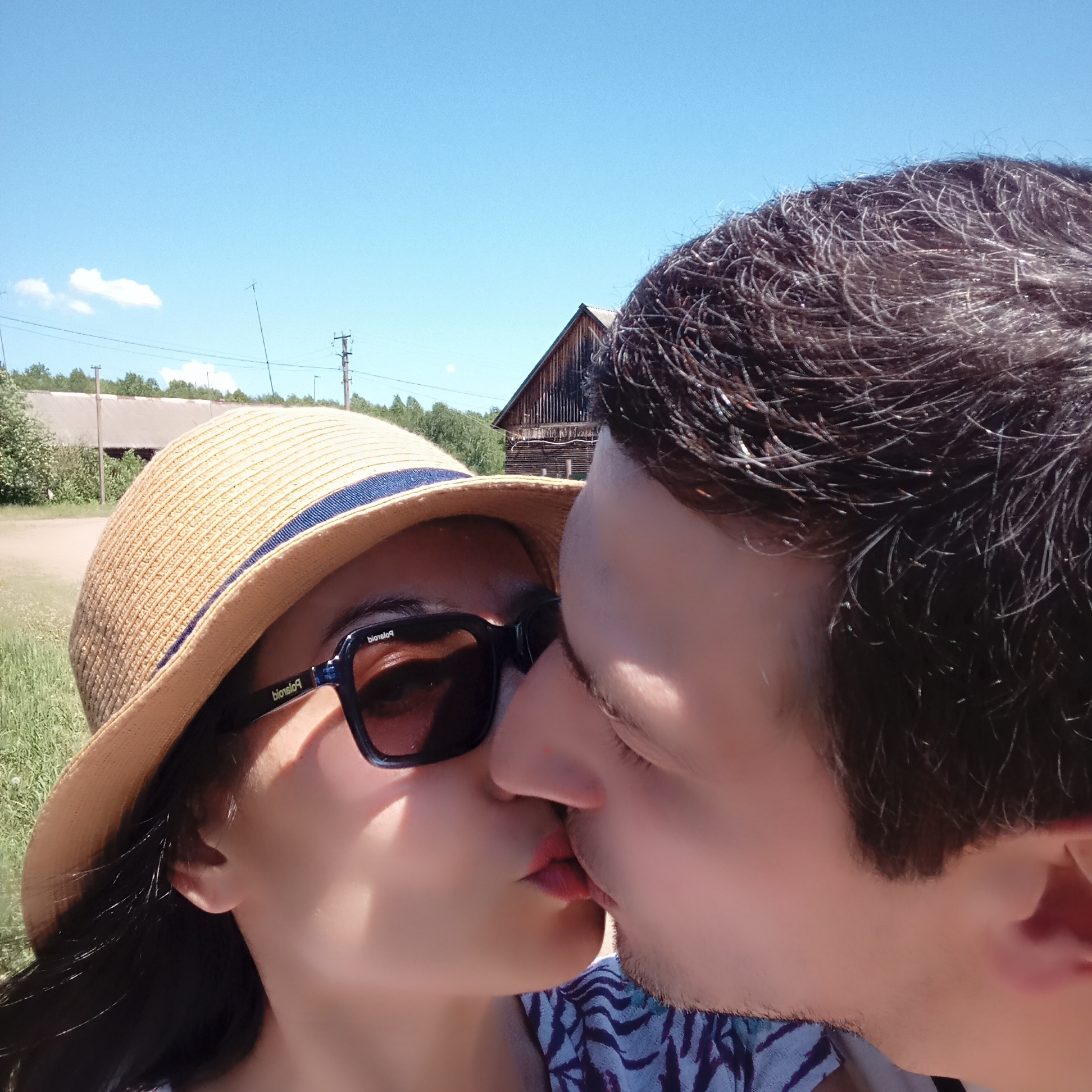 Two individuals, a man and a woman, are kissing. The woman is wearing a straw hat with a blue band and dark sunglasses. A weathered wooden structure, likely a barn, is visible in the background. The sky is clear and blue. The setting appears to be rural with grass and vegetation present. The photo was taken during daylight hours. The location is in Буренка, Russia.