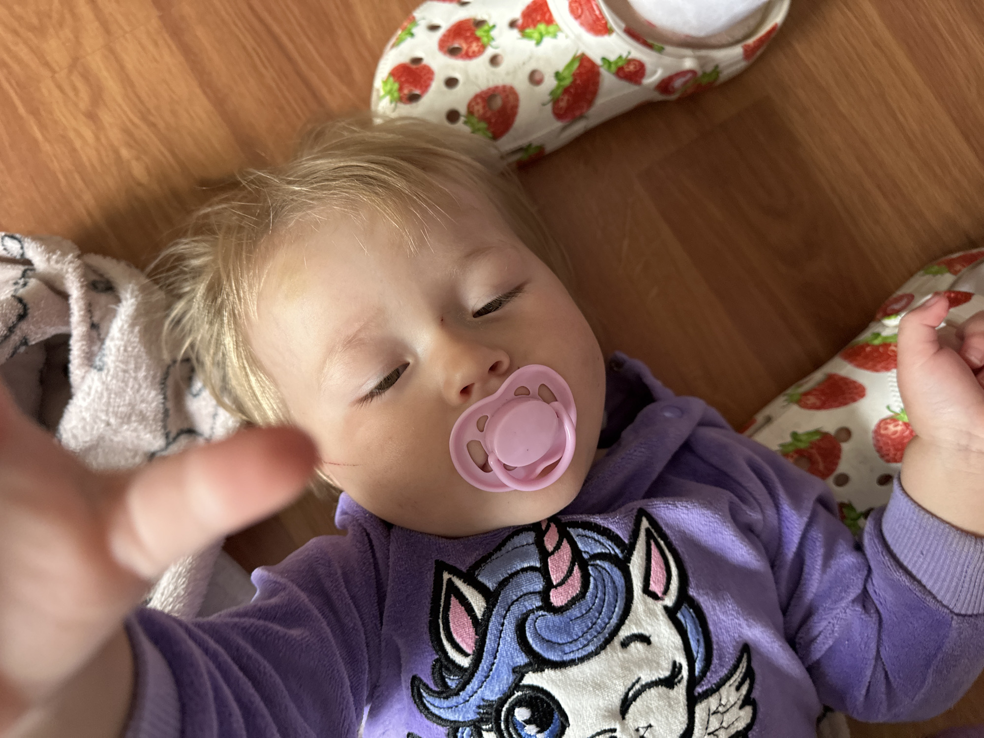 The image shows a close-up, overhead view of a baby lying on a wooden floor. The baby, with blond hair and closed eyes, has a pink pacifier in its mouth. The baby is wearing a purple hooded pajama suit with a unicorn graphic on the left side of the chest. A white sock and a white clog-style shoe decorated with strawberries are visible on the baby's right foot. A soft, light-colored blanket is partially visible behind the baby's head. The scene is not recognizable as being in Nizhny Novgorod, Russia.