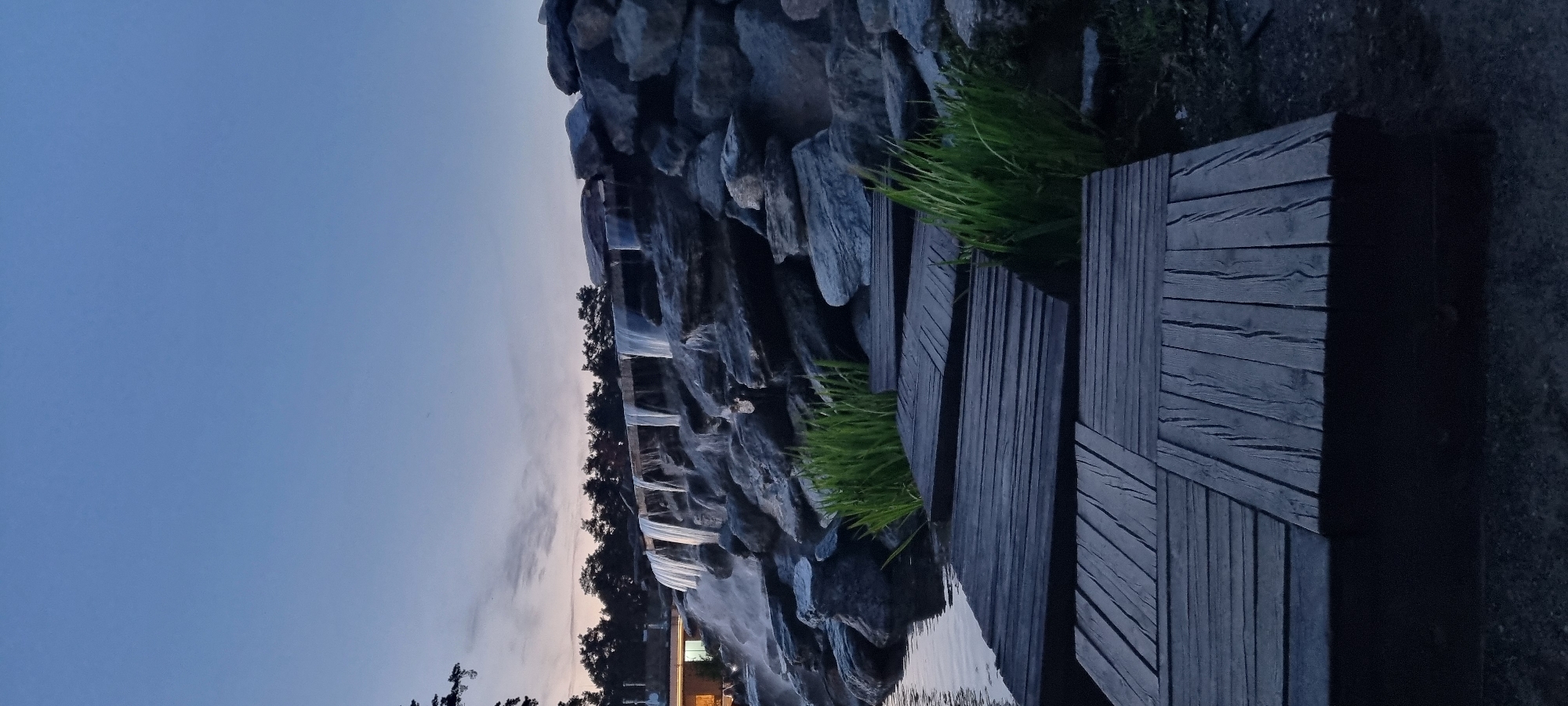 The image depicts a man-made waterfall cascading over a rock formation. In the foreground, a wooden walkway made of dark planks leads towards the water. Lush green grass grows around the rocks and between the walkway sections. The sky above is a gradient of deep blue to lighter hues near the horizon, with some faint clouds. Trees line the horizon in the background. The scene is set during dusk or early evening.