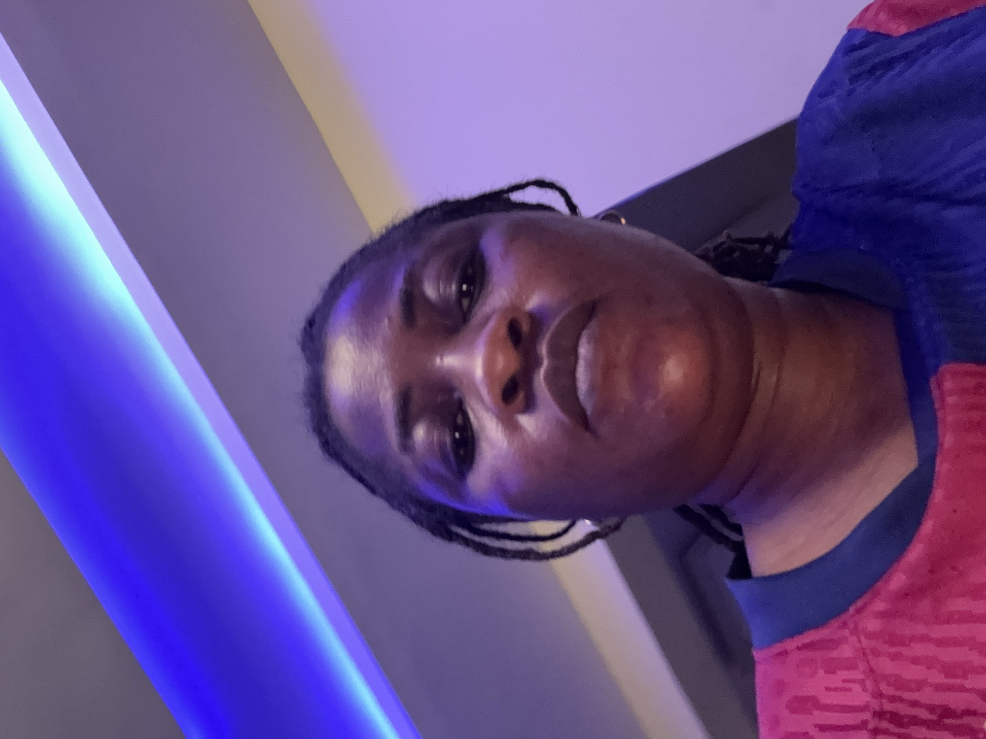 A low-angle, full-frame, medium-shot photograph captures a dark-skinned woman from the shoulders up, looking directly at the camera. She has dark hair styled in braids that fall around her face and shoulders. Her expression is neutral. She is wearing a vibrant pink shirt with a blue collar. The background consists of a ceiling with recessed blue LED lighting, casting a purple and blue hue over the scene. The lighting suggests an indoor setting, potentially a home or a public space with modern decor. No specific location in Ajara, Nigeria, can be identified from the visual information presented.