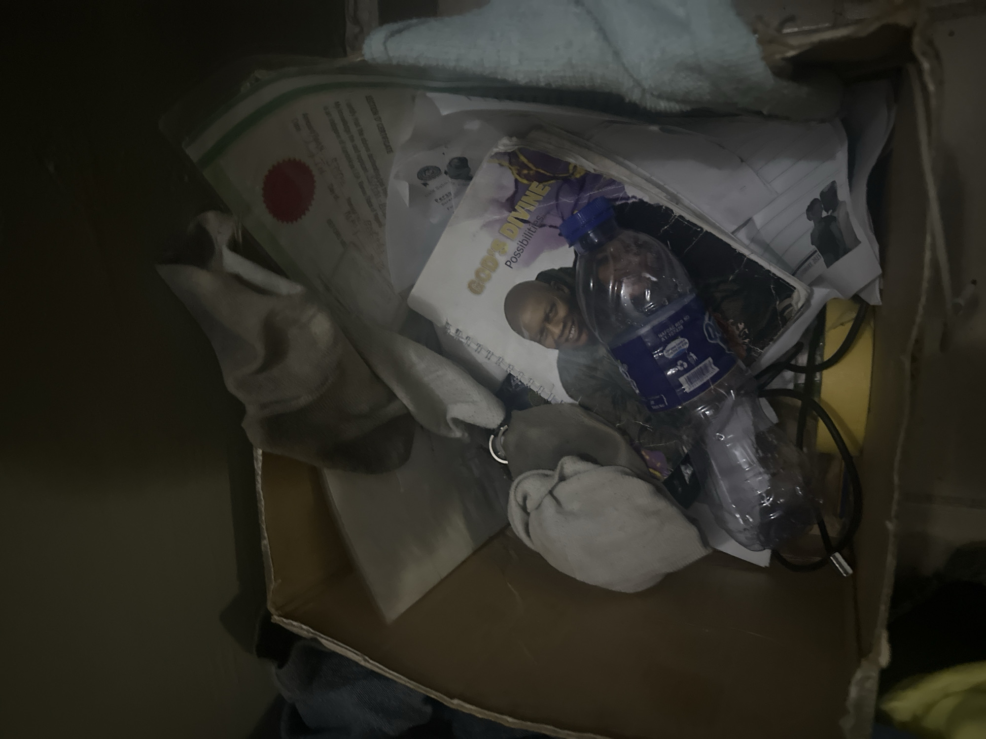 An overhead, close-up shot captures a cardboard box filled with various items. In the center of the box, a plastic water bottle with a blue cap lies on its side. Next to the bottle is a spiral-bound notebook with a picture of a smiling bald man on its cover and the words "GOD'S DIVINE Possibilities." Partially visible beneath these items are sheets of paper, one with a red circular stamp and text. A gray cloth object is also present, attached to a silver ring, possibly a keychain. Other assorted cloths and papers are scattered throughout the box, with the box itself appearing worn. The scene is dimly lit, suggesting an indoor environment.