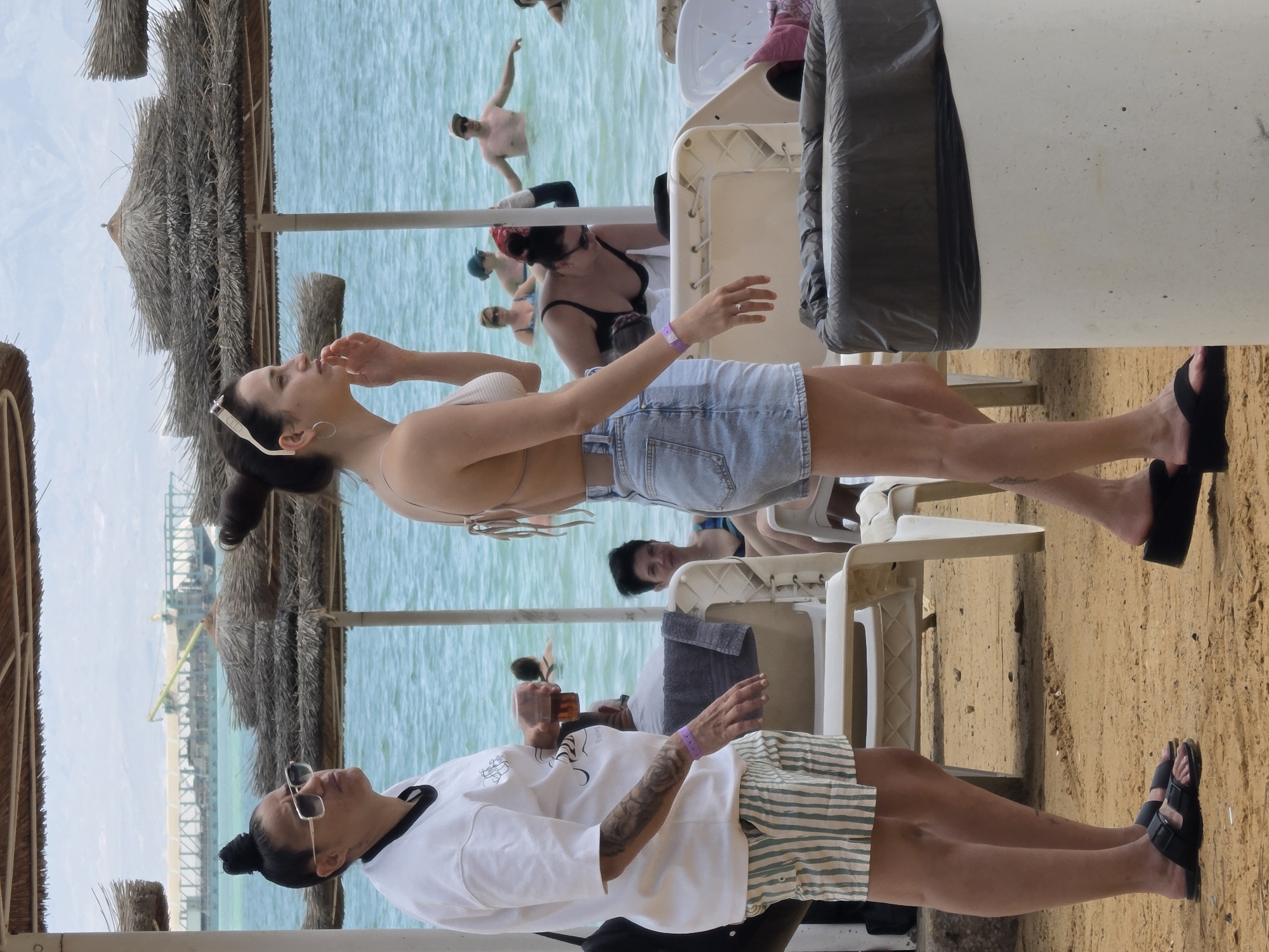 The image captures an outdoor scene at a beach in the city of Tamar Regional Council, Israel. Two women are prominently featured in the foreground, sitting on white plastic chairs. One woman is wearing a white crop top and a denim mini skirt, while the other is wearing a white t-shirt and striped shorts. Both women have dark hair and are wearing sandals.
In the background, there is a body of water with people swimming and standing. There are also thatched umbrellas providing shade. The beach has light-colored sand. In the distance, a pier or structure extends into the water. The sky is overcast. There is also a large cylindrical container next to one of the women.