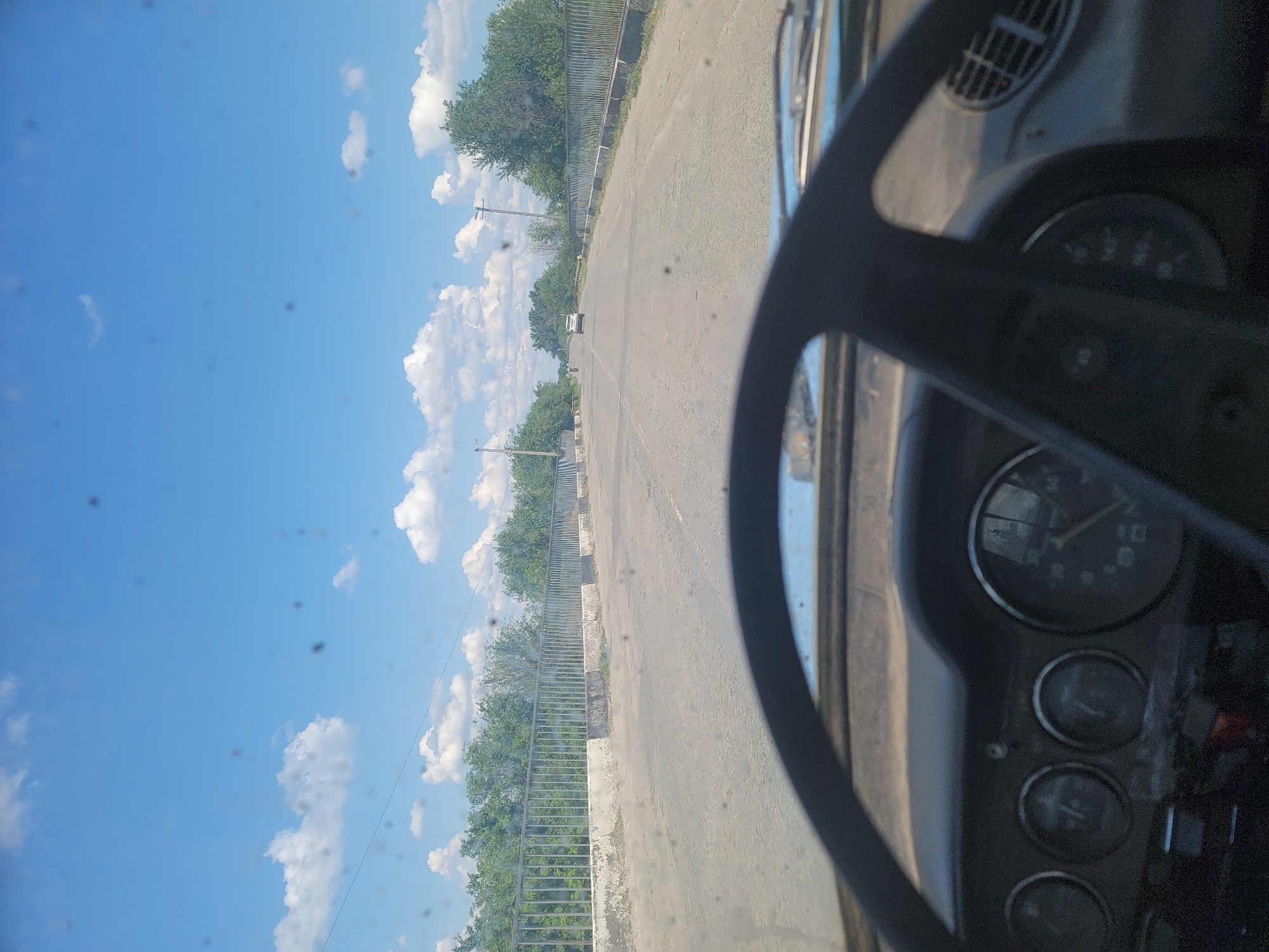 The image is taken from the driver's perspective inside a car, looking forward. The dashboard is visible in the foreground, showing several gauges, including what appears to be a speedometer. The steering wheel is also prominently displayed. The car is on a paved road, which appears to be a bridge, with metal railings on either side. Beyond the railings, there are trees and vegetation. The sky is blue with scattered white clouds. In the distance, other vehicles are visible on the road, including a white SUV and what looks like a motorcycle. The road appears to be in a rural or semi-rural setting, possibly within the city of Pereshchepyne, Ukraine. There are some dark spots on the windshield, which might be insects or debris.