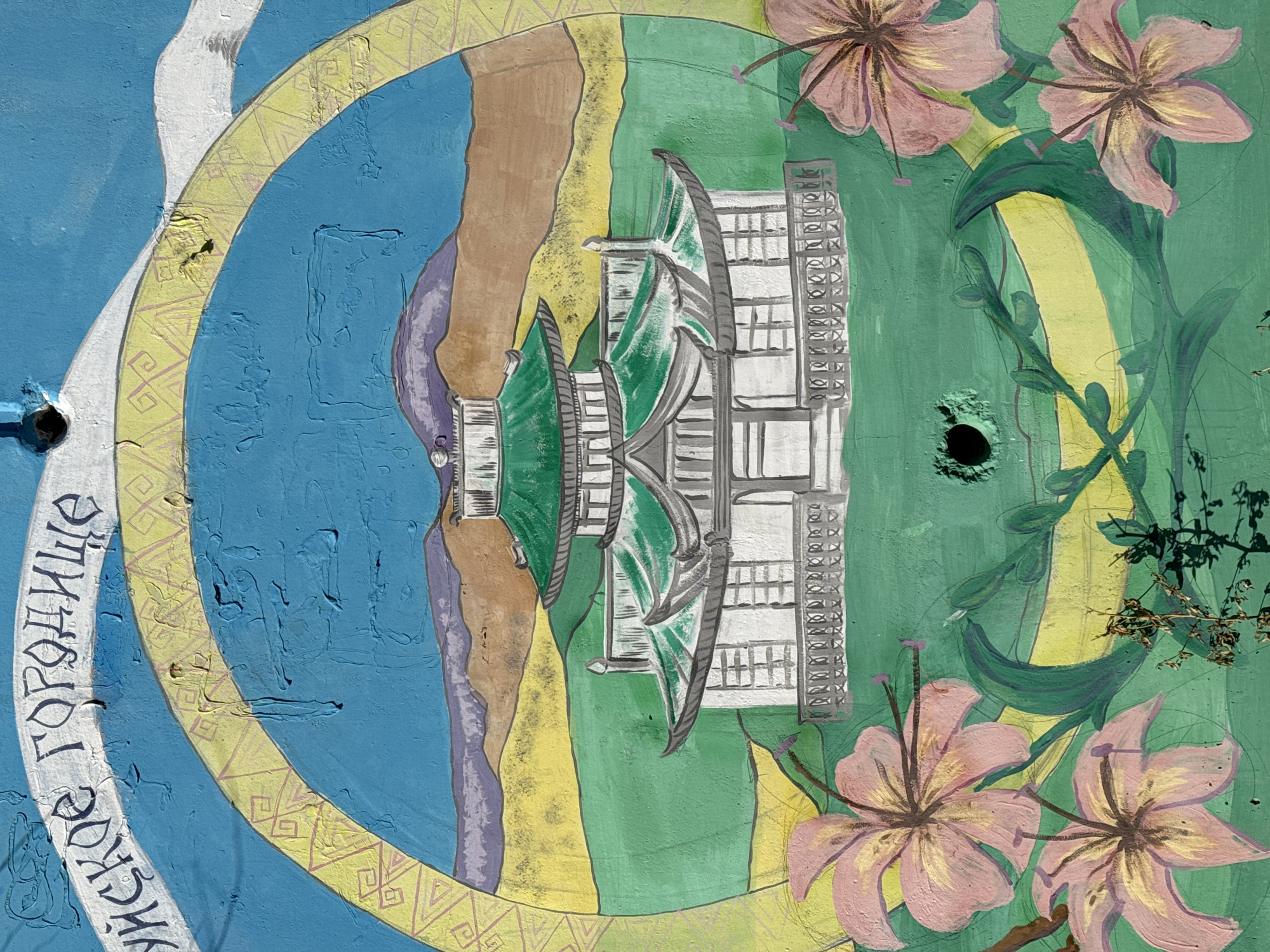 The image shows a painted mural, possibly on a wall or outdoor surface, depicting a stylized scene. The central element is a modern architectural structure, likely a building, with a green roof and gray detailing. The building appears mirrored vertically, creating a symmetrical effect. The structure is positioned within a circular composition. The background is divided into sections of blue (perhaps representing water), brown, yellow, and green. Pink flowers are painted around the right side of the circle. A yellow patterned ring surrounds the central scene. To the left, a white inscription in Cyrillic is visible. The paint texture is uneven and worn. There are holes in the surface of the wall. The location is Borzya, Russia.