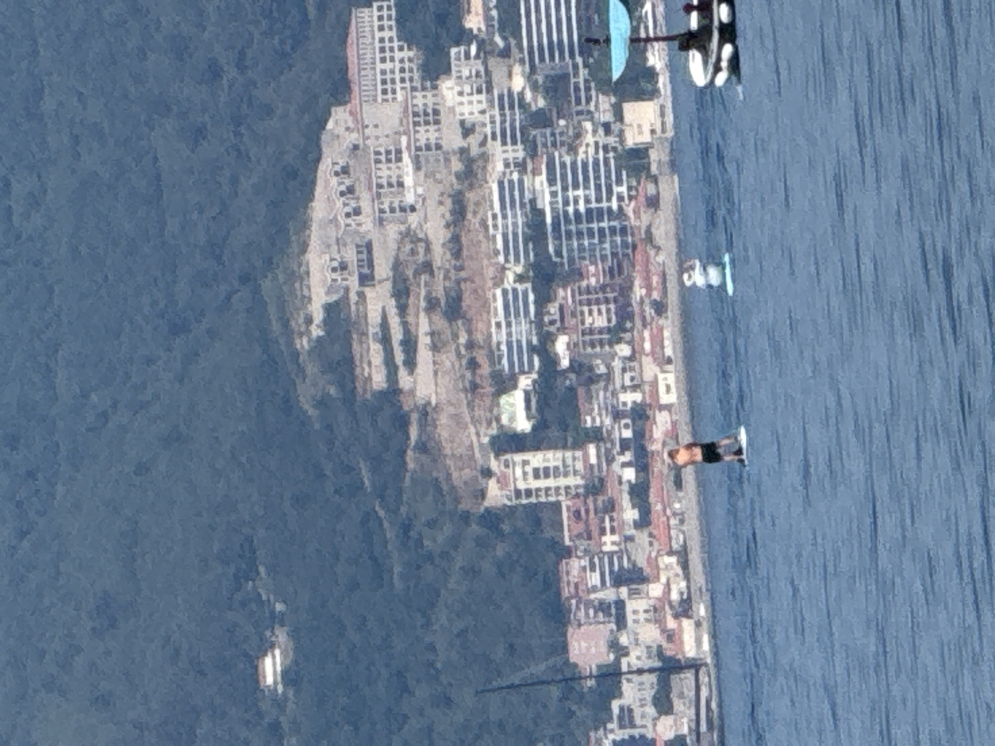 This media file captures an aerial view of a coastal town with a mountainous backdrop, likely Budva, Montenegro. The scene is dominated by the azure blue water of the sea on the right, which transitions to a densely built urban area on the left. The town features numerous buildings with white and beige facades, stacked closely together along the coastline. Many of these buildings have a distinctly Mediterranean architectural style with multiple stories and repetitive window patterns. Some buildings appear to be hotels or apartment complexes, indicated by their scale and design. Behind the urban sprawl, a lush green mountainside rises steeply, covered in dense vegetation.
In the water, a person is visible on a paddleboard, appearing to be enjoying a recreational activity. The person is wearing dark shorts and is positioned in a standing pose on the board. Nearby, a small white boat or raft is also present in the water. Further out in the sea, a few distant white structures suggest additional boats or buoys. The image's perspective is from a high vantage point, possibly from a drone or a tall building, looking down and across the landscape.
The lighting suggests it is daytime, with the sun casting shadows and illuminating the scene. The water appears calm, indicating potentially pleasant weather conditions. The clear blue sky and visible sunlight contribute to a bright and inviting atmosphere, hinting at a summer day. No visible text is present in the media file. The overall impression is one of a vibrant coastal resort town basking in the sun.