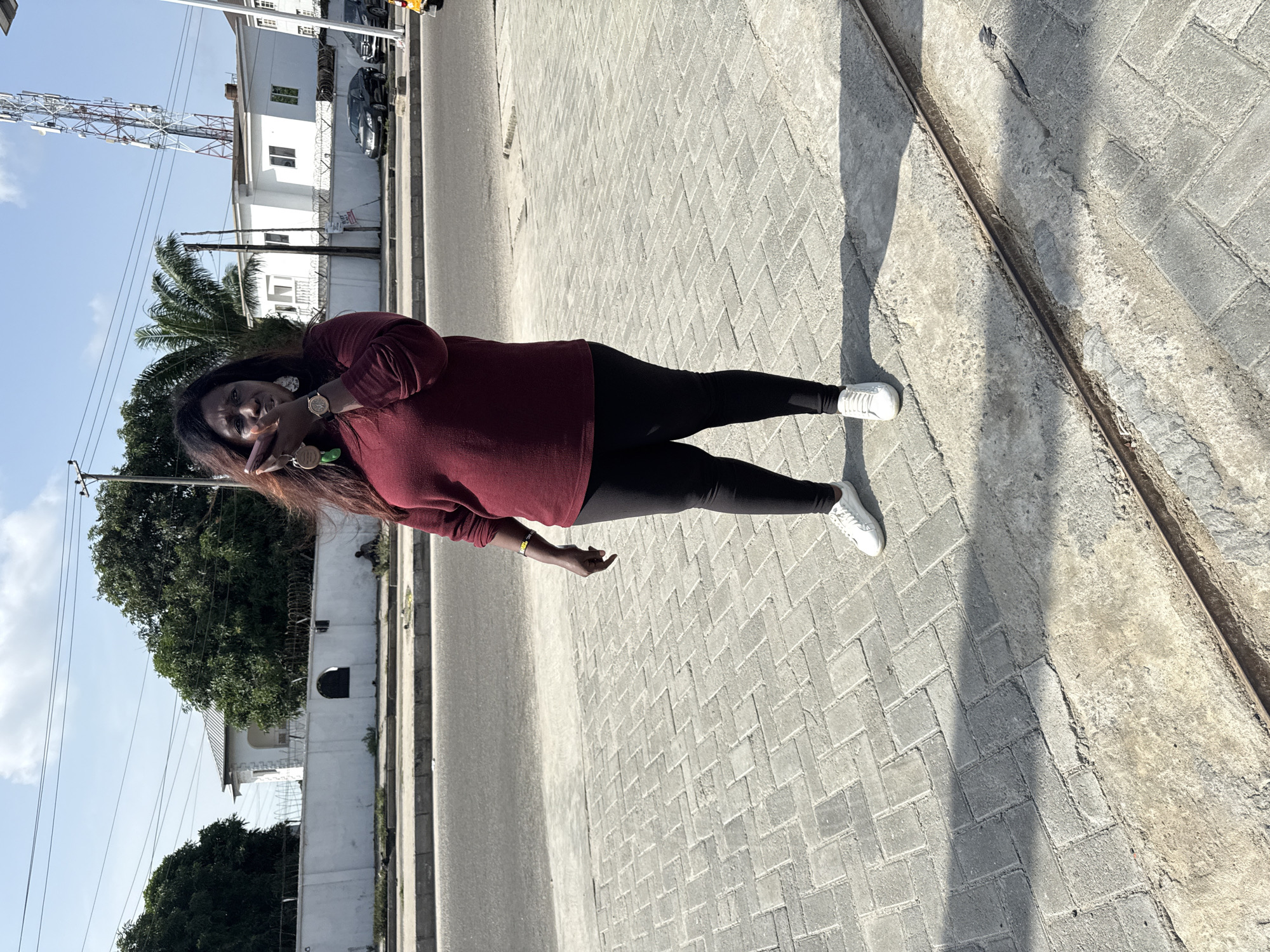A woman stands on a brick sidewalk in Ado, Nigeria. She wears a maroon sweater, black leggings, and white sneakers. Her dark hair is partially brown at the ends. She is making a peace sign with her right hand. Buildings, trees, and a cell tower are visible in the background. The sidewalk is adjacent to a road.