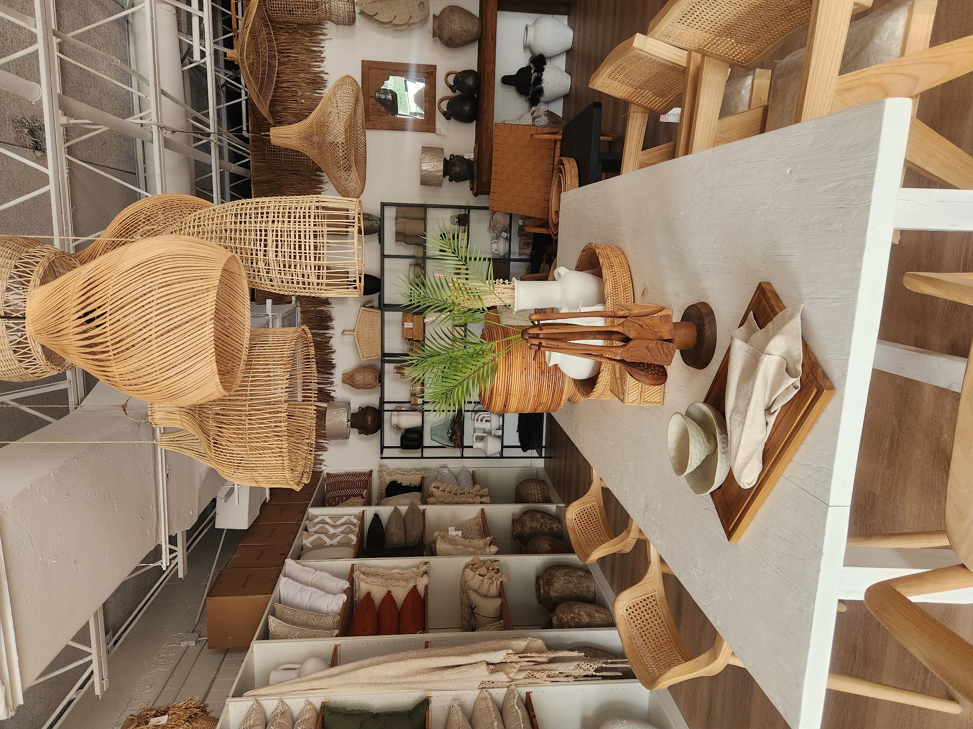 The image depicts a well-lit interior of a retail establishment, likely a home decor store, with a focus on natural materials and bohemian-inspired furnishings. The scene is set in Maitland, United States of America.
In the foreground, a long, rectangular white dining table is the central subject. The table has a textured finish and is supported by light-colored wooden legs. On the table, a wooden cutting board holds a stack of beige linen napkins and two ceramic bowls. To the left of the cutting board, another small ceramic bowl rests on the table. A wooden utensil holder with four carved wooden utensils stands to the right of the cutting board. A woven basket filled with green foliage, likely a potted plant, is positioned behind the utensils.
Surrounding the table are several light-colored wooden chairs with woven cane seats and backs. Two chairs are visible on the left side of the table, and two more are on the right.
The background reveals a retail environment. To the left, tall white shelving units are filled with an array of decorative items, including pillows in various textures and patterns, woven baskets, and large ceramic vases. A beige, fringed throw or blanket is draped over one of the shelves. To the right of the shelves, a black metal shelving unit displays more decorative objects, such as vases and lamps.
Above, a collection of large, hanging pendant lamps made from woven natural materials like rattan and wicker are suspended from the ceiling. These lamps vary in size and shape, contributing to the airy, natural aesthetic of the store. Further back, more shelving units and display areas are visible, stocked with similar decorative items. A wooden console table with two dark ceramic urns and a mirror above it can be seen against the back wall. Two large, dark urns sit on the floor in the lower right portion of the image.
The overall atmosphere is one of a curated retail space, emphasizing natural textures, neutral color palettes, and artisanal craftsmanship. The lighting appears to be a combination of overhead ambient light and possibly natural light filtering in from unseen windows. There are no people visible in the image.