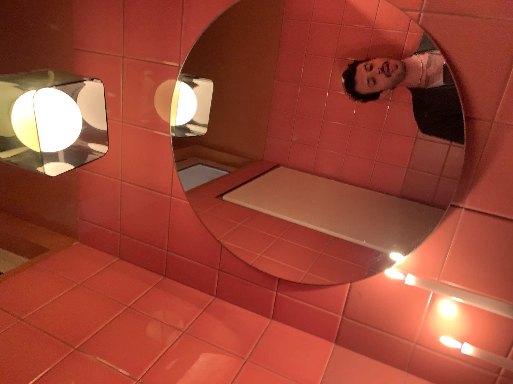 An indoor scene, rotated 90 degrees clockwise, displays a man's reflection in a large round mirror, set against a backdrop of vibrant pink-red square tiles. The man's head appears upside down in the current orientation, revealing dark, curly hair, a beard, and a playful expression as he sticks out his tongue. He is wearing a chain necklace and a dark top.
The room is uniformly tiled with the striking pink-red squares, covering both the wall where the mirror hangs and an adjacent surface, possibly a counter or floor area. To the perceived left of the mirror (which would be above it if the image were correctly oriented) is a modern light fixture. It features two glowing spherical bulbs, one larger and one smaller, enclosed within a cuboid metallic frame. This light fixture is also clearly reflected within the mirror. Further down and to the right in the image, another lighting element, appearing as a horizontal strip with multiple small, bright lights, is partially visible and also reflected.
The dominant color scheme and modern lighting suggest a uniquely designed interior space, possibly a bathroom or dressing area. The man's pose indicates a lighthearted moment, likely a mirror selfie or a casual photograph. No text is visible in the image. The location context provided is Tel-Aviv, Israel.