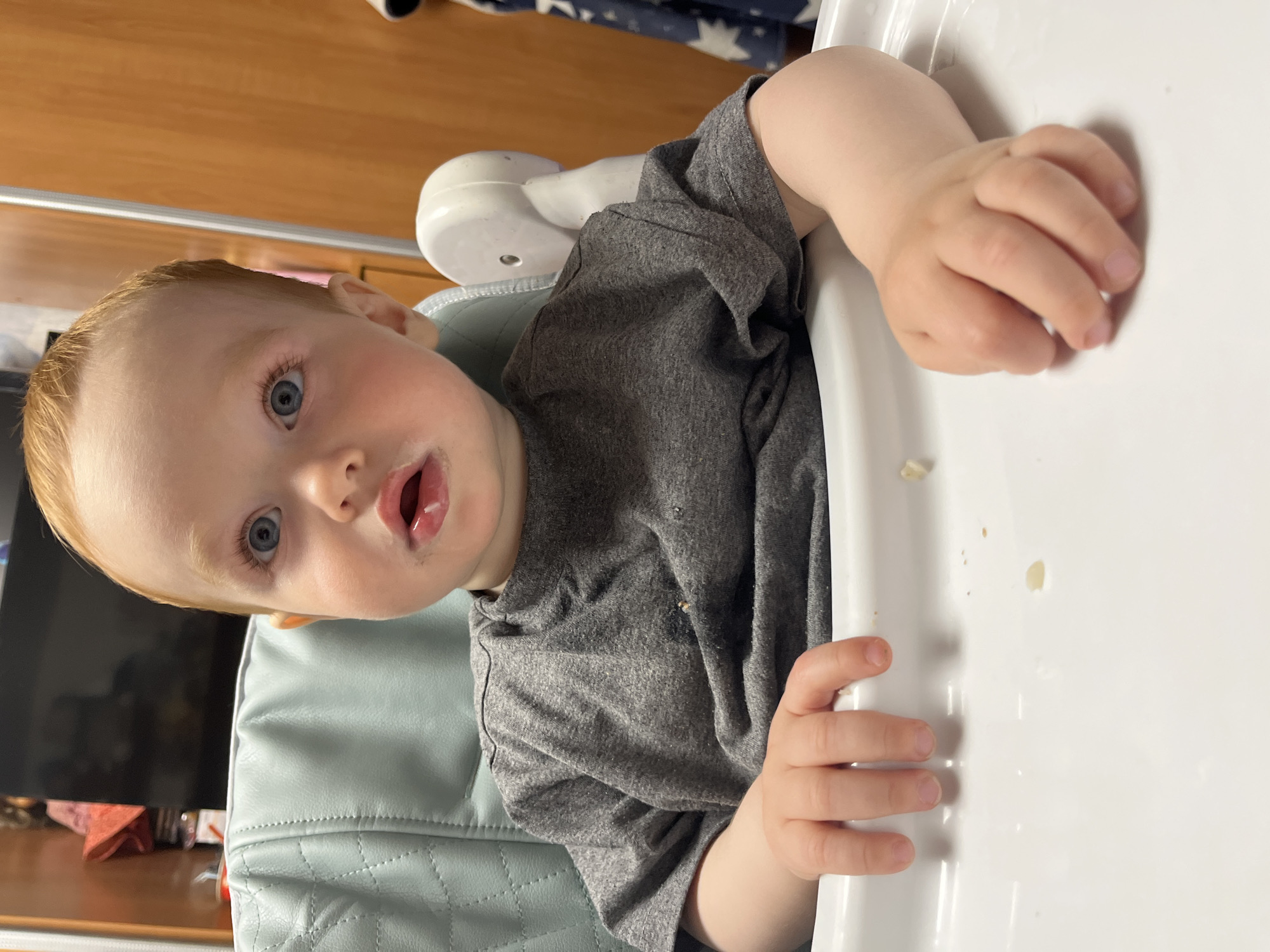 A fair-skinned toddler with light red hair and blue eyes sits in a highchair with a white tray. The toddler wears a gray t-shirt and has food residue around their mouth. The highchair has a light blue padded seat. The toddler's hands rest on the white tray of the highchair. The background includes a wooden cabinet and a glimpse of a dark-colored object, possibly a television. The image does not contain sufficient information to confirm the location as Chaikovsky, Russia.