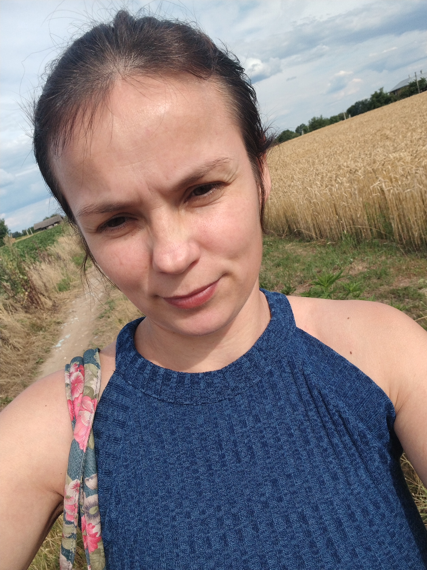 A woman is taking a selfie outdoors in a rural area, likely Molodiia, Ukraine. She has dark hair pulled back and is wearing a blue, sleeveless, ribbed top. A floral patterned strap is visible over her left shoulder. Behind her, a field of golden wheat stretches towards the horizon under a cloudy sky. A dirt path is visible to her left, with dry grass and some greenery along its edge. In the distance, some houses and trees can be seen. The overall scene suggests a sunny day in a countryside setting.