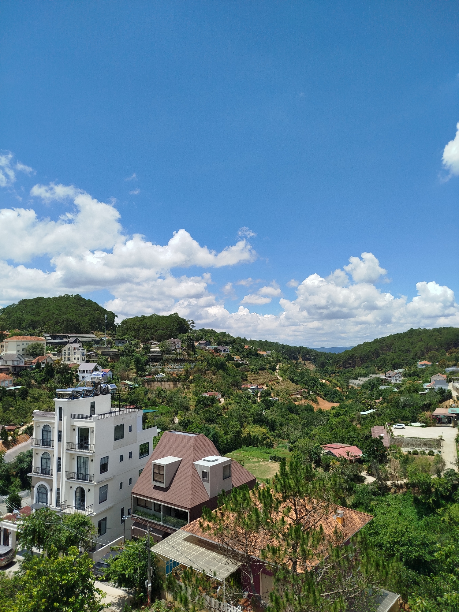 A wide-angle shot captures a residential area nestled amidst rolling hills and lush greenery under a bright, cloud-scattered blue sky. The architecture features a mix of traditional and modern styles, including multi-story buildings with balconies and red-tiled roofs. The scene suggests a high-altitude location, and based on the architecture and landscape, the image likely depicts an area in Da Lat, Vietnam. The foliage is dense, with a variety of trees and shrubs filling the valleys and slopes.
