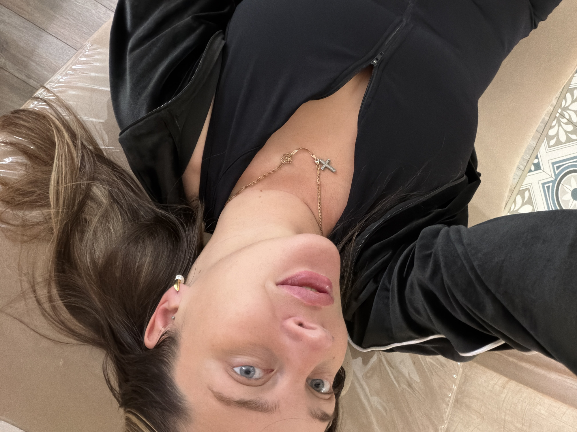 A woman lies on her back. She has light skin, light blue eyes, and light brown hair with some blonde highlights. She is wearing a black zip-up jacket and a gold necklace with a cross pendant. She is wearing a gold earring in her left ear and a stud in her cartilage piercing. She is lying on what appears to be a beige surface covered with a clear plastic sheet. The floor is visible in the upper left corner, showing gray wood-look planks, and there is a decorative tiled area in the upper right corner. The location is Unknown, Unknown.
