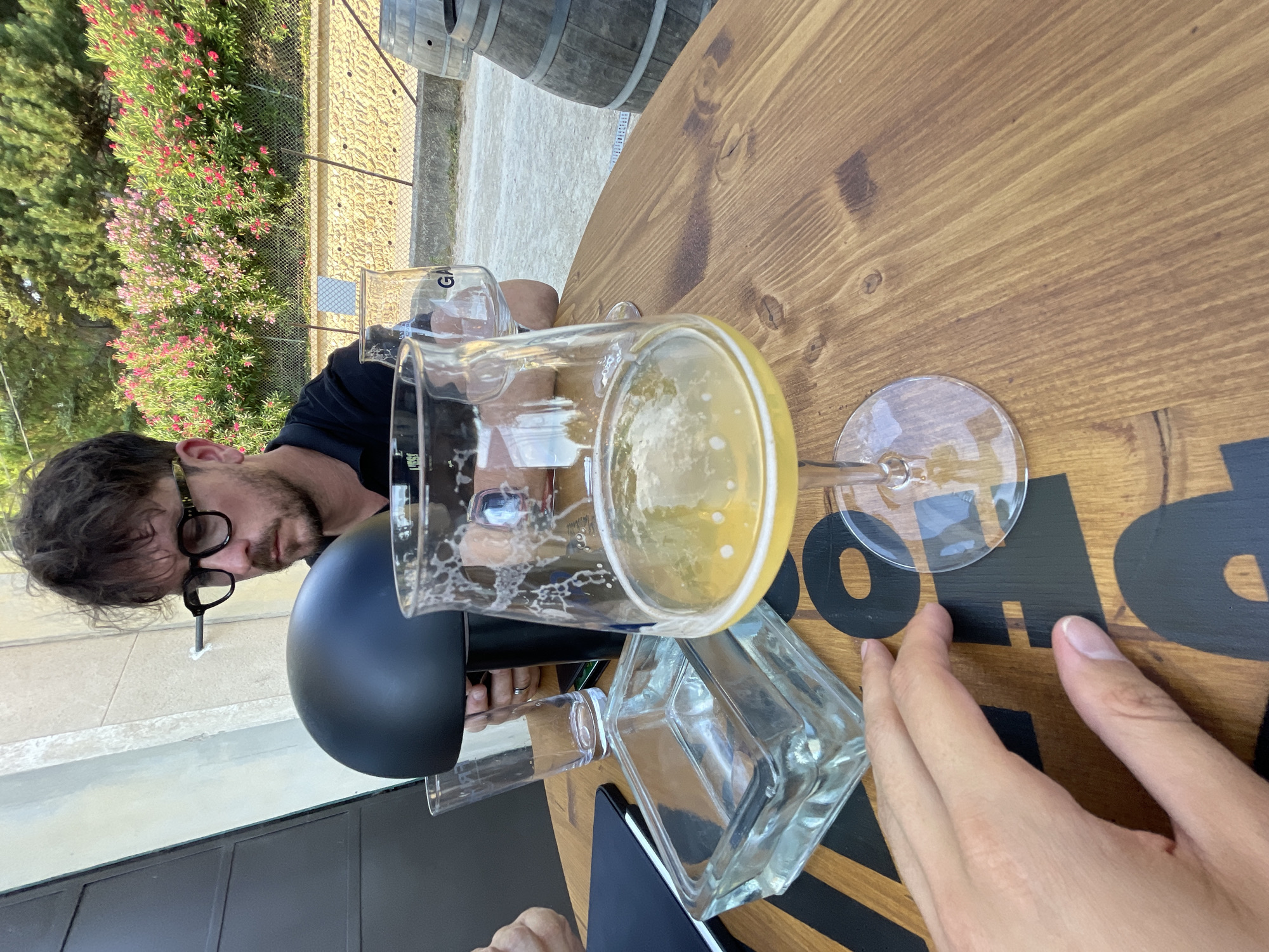 In Cucuron, France, at an outdoor table with a wooden surface, a man with dark, curly hair, a beard, and glasses sits and looks down. He is wearing a black polo shirt. In front of him, a beer glass sits partially filled with a light-colored beverage, with foam on its rim and sides. An empty glass is visible behind the beer glass, and another glass is further back. To the left of the beer glass, a rectangular glass ashtray sits empty. A dark notebook or folder is also on the table, partially obscured by a person's hand reaching into the frame from the bottom left. In the background, a fence separates the seating area from a path or road, beyond which is a stone wall and lush greenery with pink flowers. Two wooden barrels are visible to the right.