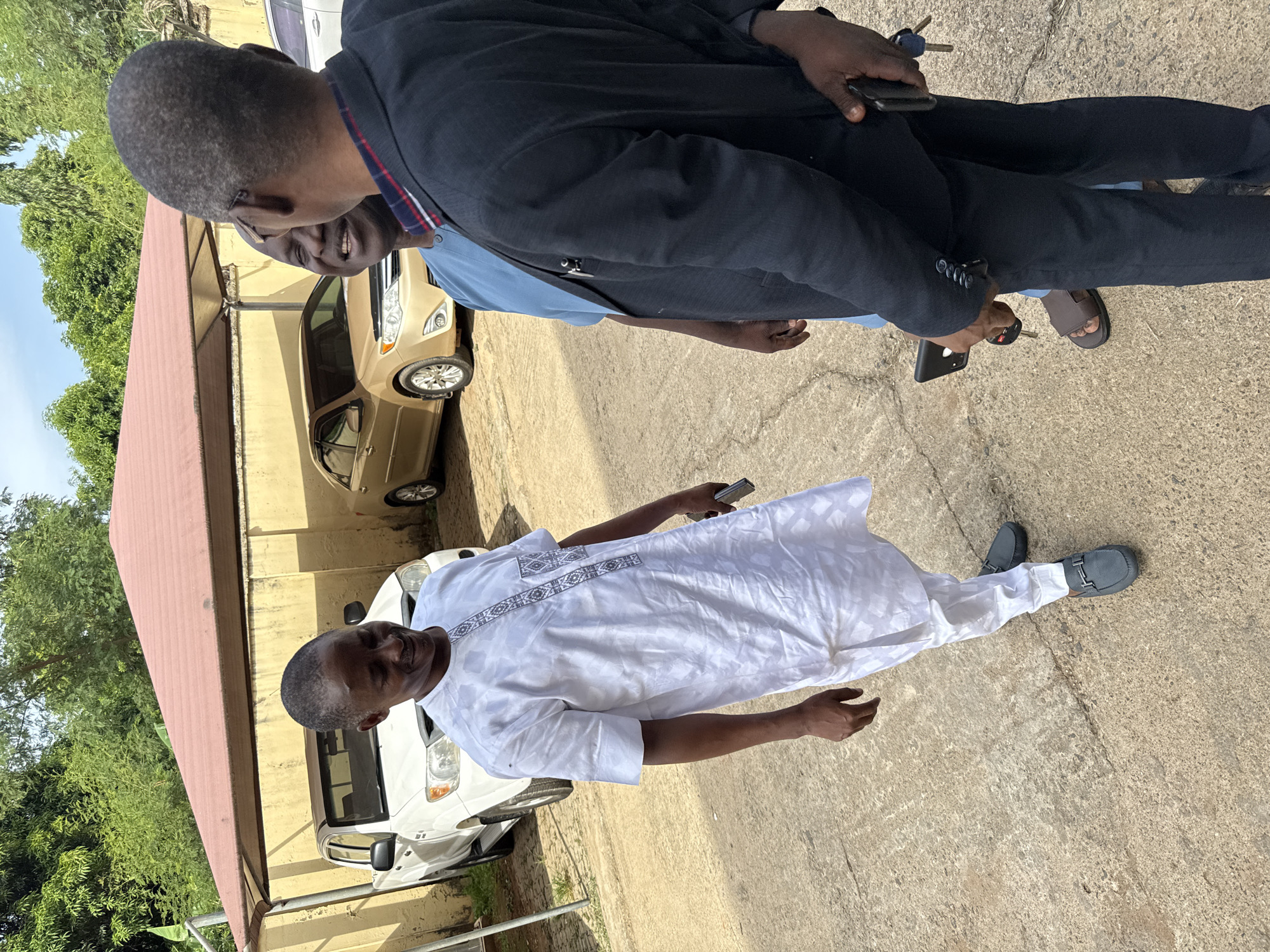 The image captures an outdoor scene featuring three men on a sunny day in what appears to be a paved courtyard or parking area, consistent with a setting in Abuja, Nigeria.
In the foreground, a man with a dark complexion and closely cropped hair stands, looking towards the viewer or slightly to the side. He is dressed in a white traditional Nigerian outfit, possibly an agbada or similar attire, adorned with intricate grey and black embroidery across the chest and down the front. He wears dark grey loafers and holds a black smartphone in his right hand.
Behind him, two other men are partially visible, engaged in a conversation. The man closest to the foreground, positioned sideways, has a dark complexion, short hair, and is wearing a light blue collared shirt. He appears to be smiling. The third man, mostly seen from behind and to the left of the second man, wears a dark suit jacket over a light blue shirt with red and white striped detailing on the collar. He holds a set of keys and a smartphone in his right hand and is wearing brown sandals.
The background includes a golden-colored sedan, resembling a Toyota Camry, parked on the left, and a white vehicle further back. A brown corrugated roof structure or canopy extends over part of the area on the left. Beyond the roof and vehicles, lush green trees and a yellow-walled building are visible under a bright blue sky. The ground is a light-colored concrete or asphalt pavement. The strong sunlight casts clear shadows, indicating bright daytime conditions. No discernible text is visible in the image.