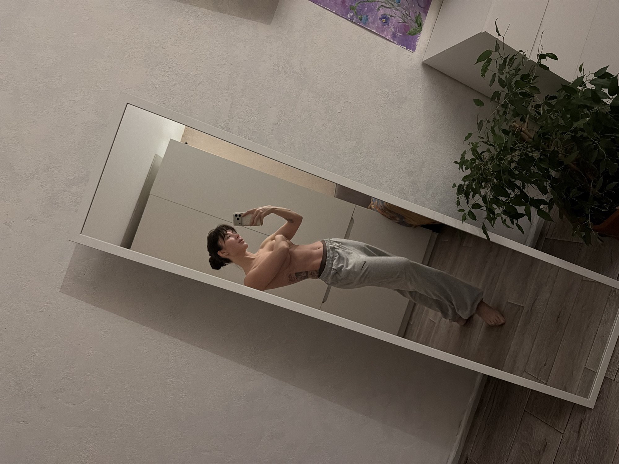The image, taken in Perm, Russia, captures a person taking a mirror selfie indoors. The scene is significantly tilted, giving it a dynamic angle.
The main subject is a person, appearing to be female, reflected in a large, white-framed full-length mirror. She is holding a light-colored smartphone (possibly an iPhone) up to take the photo. Her dark hair is styled in a bun. She is shirtless, showcasing a toned physique, and wears light grey sweatpants with a "Nike" logo visible on the left thigh. A tattoo is visible on her right arm and side of her torso. Her expression is neutral, focused on the act of taking the picture.
The mirror is positioned against a light-colored, textured wall. Reflected in the mirror and visible around it are elements of an indoor setting: a light wooden floor, white cabinet or wardrobe doors, and a potted green leafy plant. Above the mirror's reflection, on the real wall, a small abstract painting with purple tones is partially visible. The lighting is artificial and somewhat dim, suggesting it might be evening or night, or a room with limited natural light.