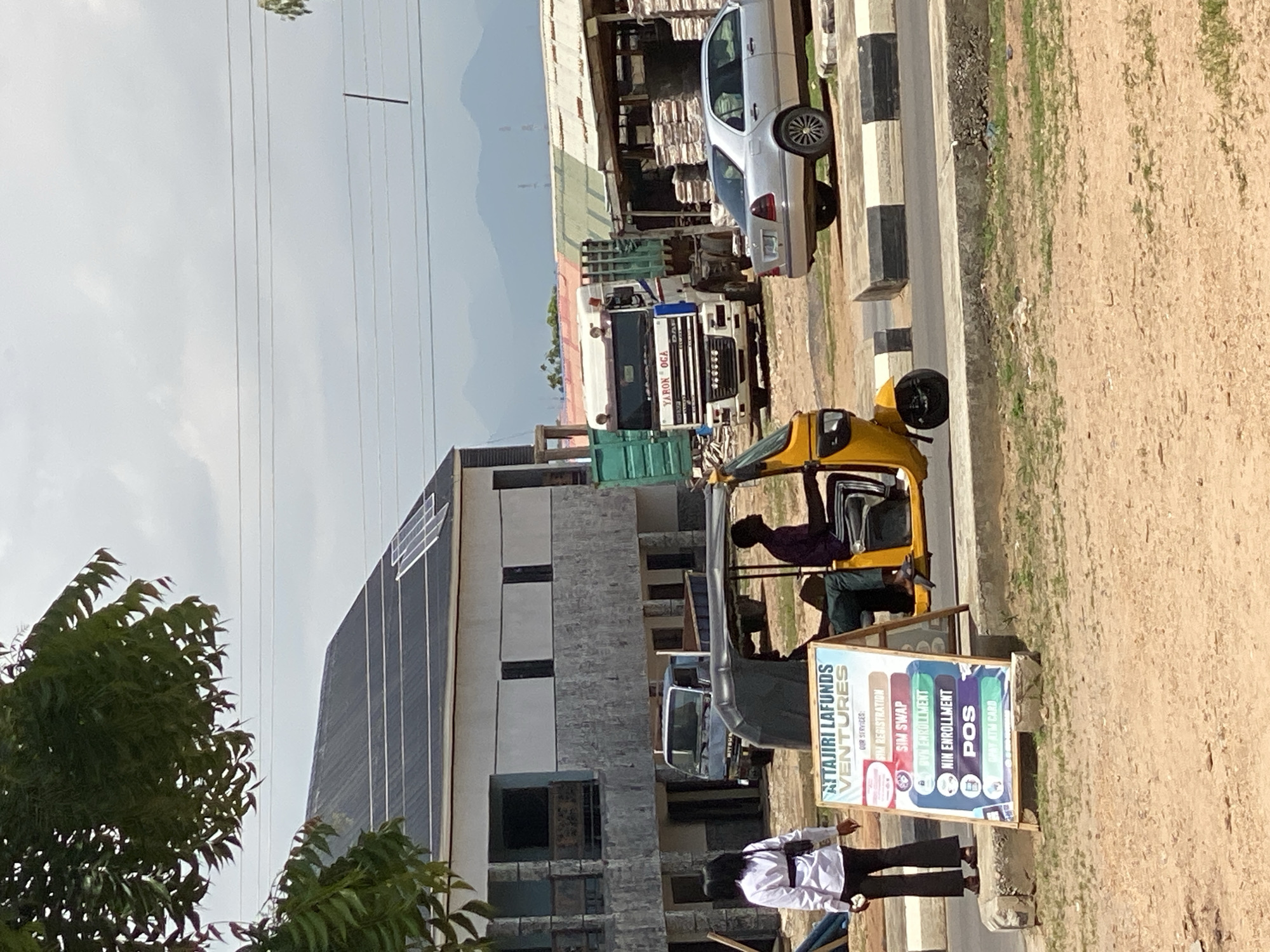 This media file captures a street scene in Jalingo, Nigeria, likely during daylight hours with overcast skies. The setting appears to be a commercial or industrial area, with buildings under construction or renovation, and parked vehicles lining the street.
Several subjects are visible. In the foreground, two people are standing near a roadside business. One is wearing a white shirt and black pants, while the other is in dark clothing. A yellow, three-wheeled auto-rickshaw, also known as a "Keke," is parked prominently in the mid-ground. A person is seated in the driver's position of the auto-rickshaw, and another person is partially visible in the passenger area. Further down the street, a large truck with a green container is parked, and behind it, a silver car is visible.
The activity taking place seems to be the day-to-day operations of a street-side business. The auto-rickshaw and its occupants, along with the people standing nearby, suggest potential customer interaction or waiting. The construction of the building in the background implies ongoing development in the area.
Notable details include the presence of solar panels on the roof of one of the buildings, indicating a modern adaptation. A colorful A-frame sign stands near the auto-rickshaw, displaying text. The visible text on the sign reads: "TAJIRI LAfunds VENTURES," and lists services such as "SIM REGISTRATION," "SIM SWAP," "NIN ENROLLMENT," and "POS." This suggests the business offers mobile network and identification-related services, as well as point-of-sale transactions. The overall atmosphere seems calm and functional, typical of a busy urban environment.