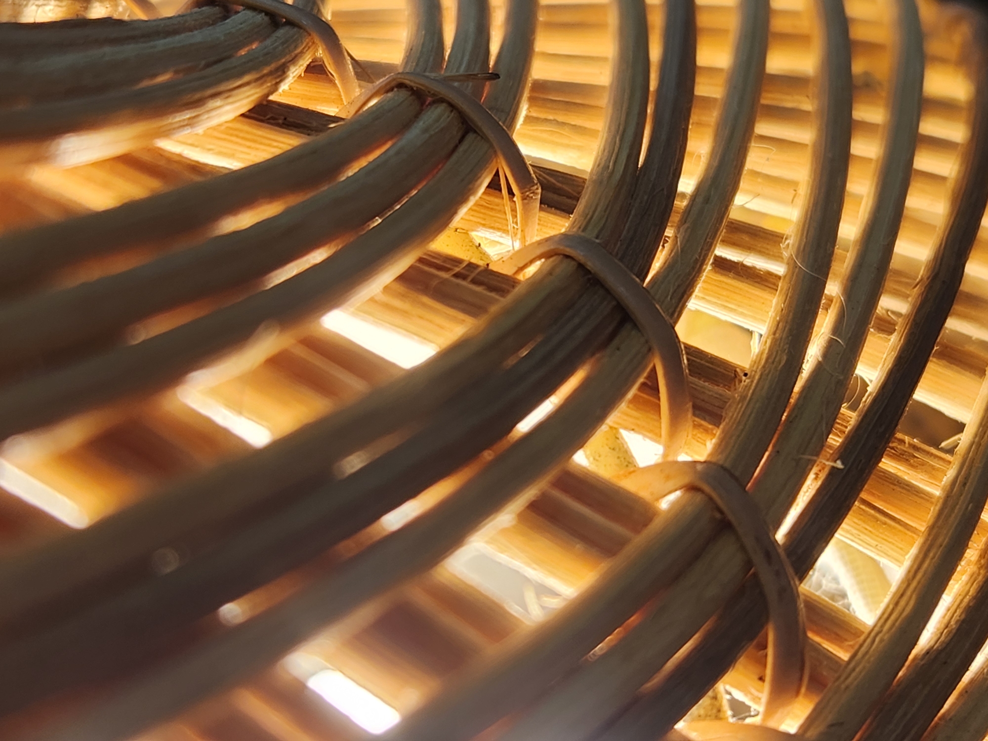 This is an extreme close-up, high-angle shot of a woven basket. The basket is made of natural, light-brown wicker or rattan material, with individual strands appearing to be about 0.5 to 1 centimeter in diameter. The weaving creates a crisscross pattern, with some strands running horizontally and others diagonally. The light source appears to be behind and to the left of the basket, creating bright highlights and casting shadows within the woven structure. The lighting gives the basket a warm, golden-orange glow. No people, animals, or other objects are visible in the frame. The image appears to be taken indoors with soft, diffused lighting. There is no information in the image to suggest a specific location in Maitland, United States of America.