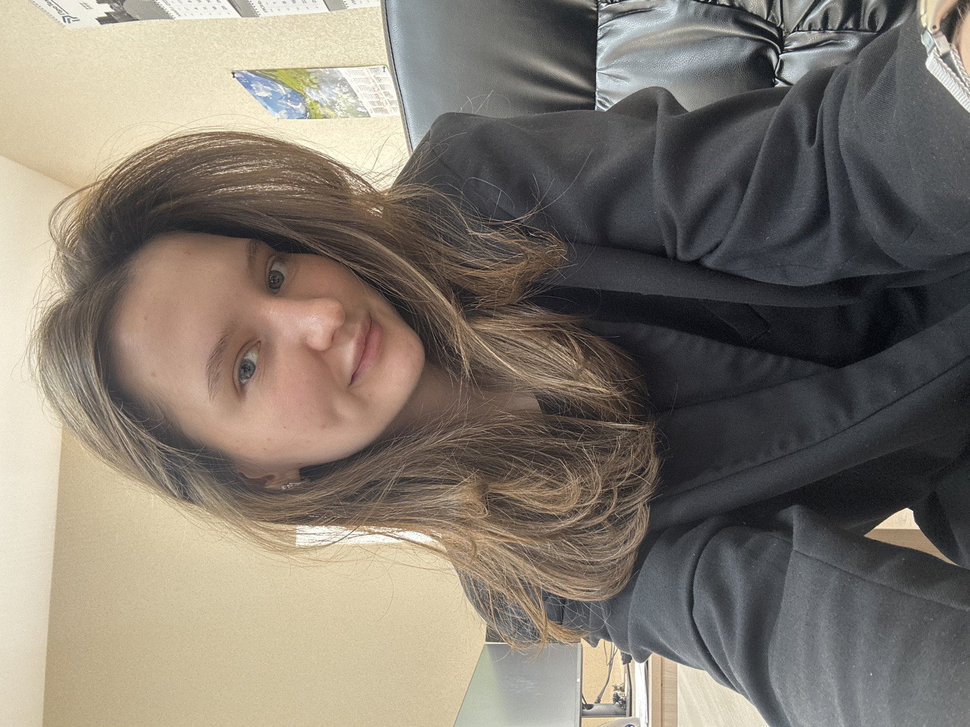 A young woman is featured in a selfie, taken indoors in what appears to be an office or home office setting in Perm, Russia. She is shown from the chest up, with her head slightly tilted and a gentle, neutral expression, almost a faint smile, as she looks directly at the camera. Her long, light brown hair, with subtle highlights, is parted on the side and cascades around her shoulders. She has light-colored eyes, possibly blue or green.
The woman is wearing a dark, likely black, blazer or jacket over a dark top. Behind her, a black faux leather office chair is partially visible. The background consists of a plain, light beige or cream-colored wall. On the wall, two items are hung: one is a paper with some text and numbers, possibly a calendar or chart, and below it, another paper featuring a landscape image, also likely a calendar page. A desk or table surface is visible in the lower portion of the frame, along with a glimpse of what might be a computer monitor or equipment. The overall lighting suggests an indoor setting during the daytime. The text visible on the papers on the wall is not clearly legible.