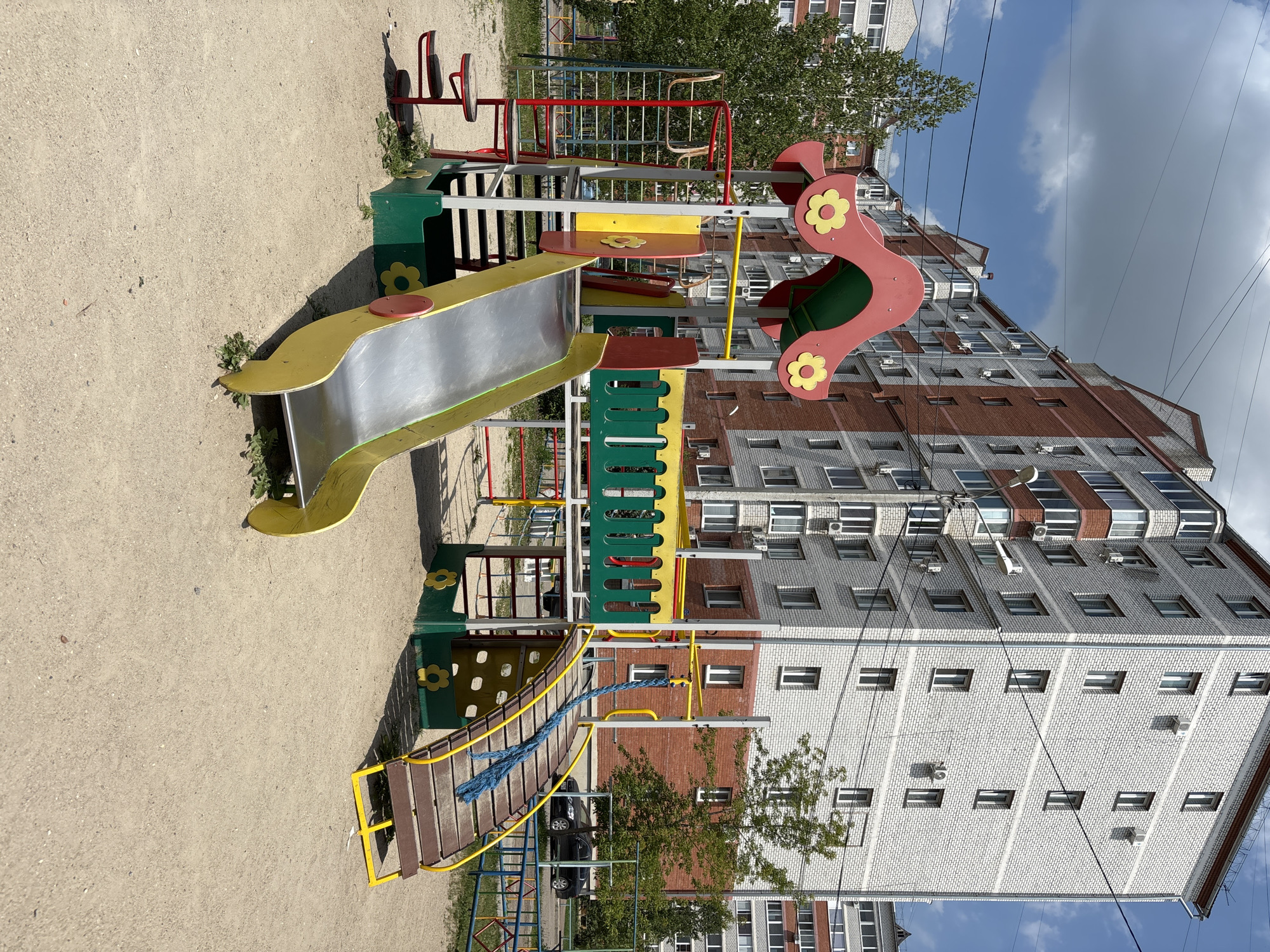 An outdoor playground with a multi-colored slide, climbing structure, and rope bridge is situated in a sandy area. In the background, tall brick apartment buildings with numerous windows and air conditioning units are visible under a partially cloudy sky. A black car is parked on the street behind a low fence. This scene is located in the city of Blagoveshchensk, Russia.