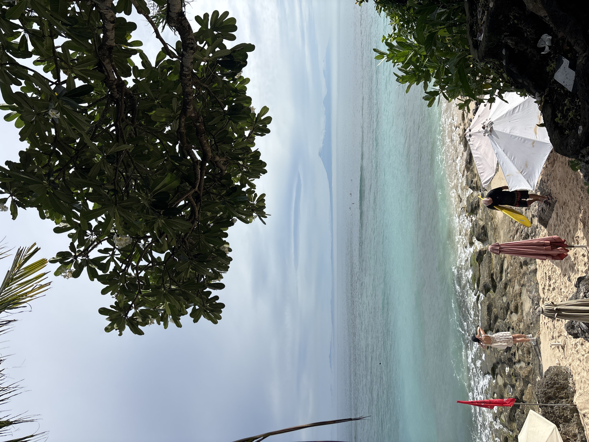 The image depicts a beach scene in Kuta Selatan, Indonesia. The foreground contains tree branches on the left and a rocky outcrop with vegetation on the right. The beach is visible below the trees. The ocean is turquoise in color, with a hazy horizon visible in the distance.
On the beach, a white umbrella provides shade for a man wearing dark clothing and holding a yellow object. Nearby, an open peach-colored umbrella is positioned near the shoreline. A person dressed in light clothing is standing on the rocks near the water's edge. A red flag is planted in the sand near the bottom of the frame.