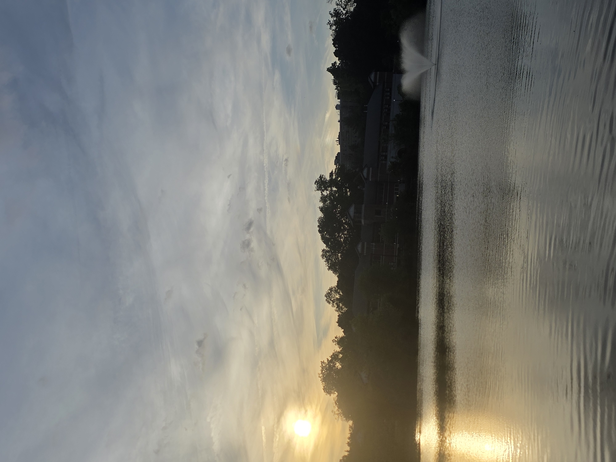 The image depicts a sunset scene over a body of water in New Rochelle, United States of America. The sun is positioned low in the sky, casting a golden glow on the water and surrounding landscape. Silhouetted trees and buildings line the shore. A fountain is visible in the water. The sky exhibits a mix of clouds and clear patches.