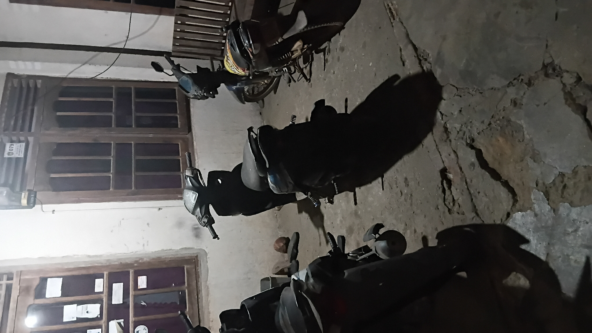The image depicts an outdoor scene at night in Prabumulih, Indonesia, likely a residential area or a small business. Three motorcycles are parked in front of a building with traditional wooden windows and a textured, possibly concrete, floor. The building appears to be a house or shop, with windows showing an interior that is not clearly discernible due to the lighting conditions. One motorcycle, a dark scooter, is parked in the foreground to the left, partially obscured. In the middle ground, another dark scooter is parked, casting a long shadow. To the right, a third motorcycle, possibly a sport bike, is parked, with a helmet resting on its seat. The ground surface is uneven and cracked, indicating age or wear. A small pile of what appears to be a pair of shoes and a coconut shell is visible near the base of the middle motorcycle. The overall ambiance is dark and quiet, suggesting it is late at night. There are no people visible in the frame.