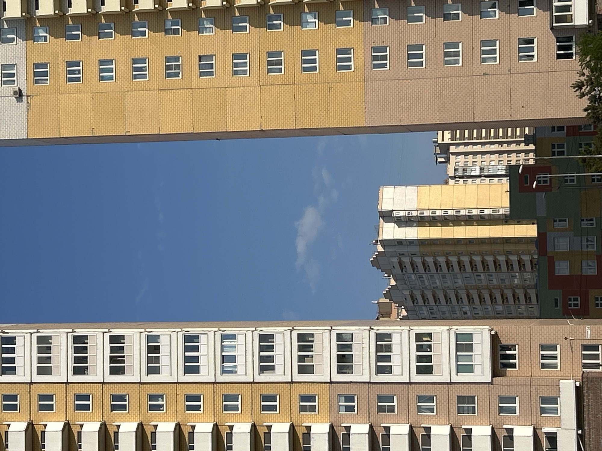 The image shows a view of apartment buildings in Saint Petersburg, Russia, shot from a low angle looking upwards. Two high-rise buildings frame a blue sky. The buildings are characterized by their blocky, modular design, with rows of windows. The buildings have a yellow-brown brick exterior. Another building is seen in the background, featuring a more complex, multi-colored facade. The sky is clear with some wispy clouds.