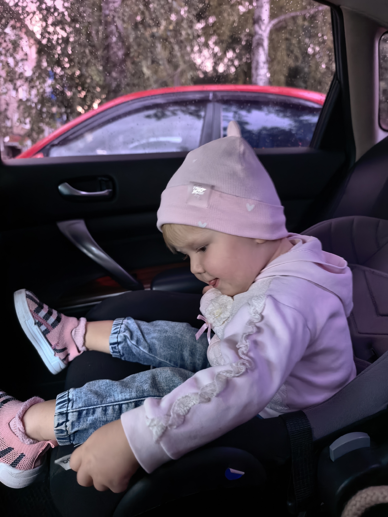 A young child, likely female, is seated in a car seat within a vehicle. The child is wearing a pink beanie, a light pink hooded jacket adorned with lace trim, blue jeans, and pink and black striped sneakers with pink ankle socks. The child is looking downwards, with their tongue slightly protruding. The car's interior is visible, including a black door handle and a dark-colored car seat. Outside the car, through the window, a red car is visible, along with trees and foliage, suggesting an outdoor setting. The weather appears to be overcast or rainy, as indicated by droplets on the window. The scene is consistent with being inside a vehicle parked or moving within the city of Sterlitamak, Russia, given the context provided.