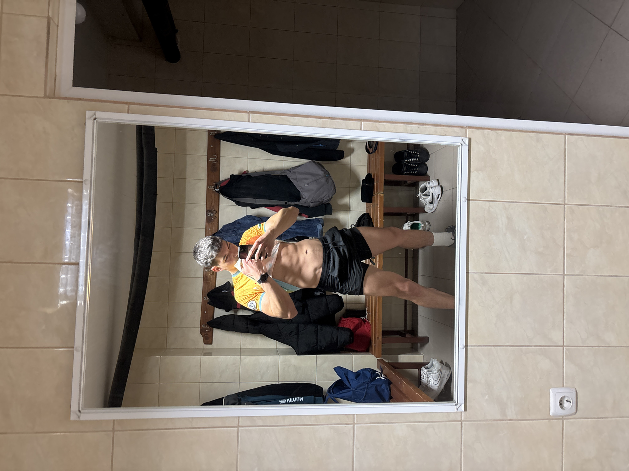 The image depicts a person taking a selfie in a mirror. The person is wearing a yellow shirt, black shorts, and white socks. They are holding a phone in their hand. The reflection in the mirror shows a hallway with various objects, including clothes hanging on hooks, shoes on a shelf, and bags on the floor. The walls of the hallway are tiled. There is an electrical outlet visible on the tiled wall. The location appears to be indoors, possibly in a building in Zlatograd, Bulgaria.
