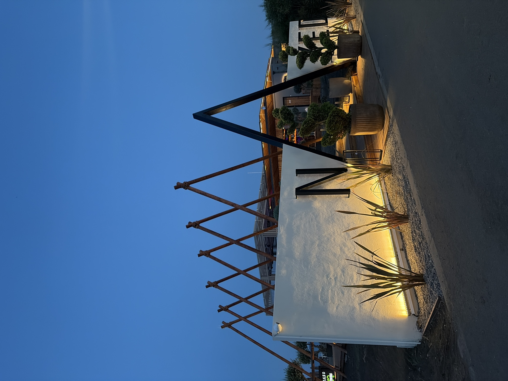 The image depicts the exterior of a modern establishment at dusk, featuring a white textured wall with the prominent black letter "N" and the partial word "ED" on its surface. Behind the wall, a building with a wooden slatted roof and an outdoor seating area is visible. Several triangular wooden structures with ropes at their apexes are erected in front of the building, creating a distinctive architectural element. Potted plants, including topiaries and tall grasses, are strategically placed around the entrance, illuminated by uplighting from a strip of lights along the base of the wall. A paved pathway or road runs in the foreground. The sky is a deep blue, indicating twilight. On the far left, a green neon sign that reads "NAKED" is partially visible. The scene appears to be located in Burgas, Bulgaria, a coastal city known for its modern architecture and seaside establishments. There are no people visible in the image.
