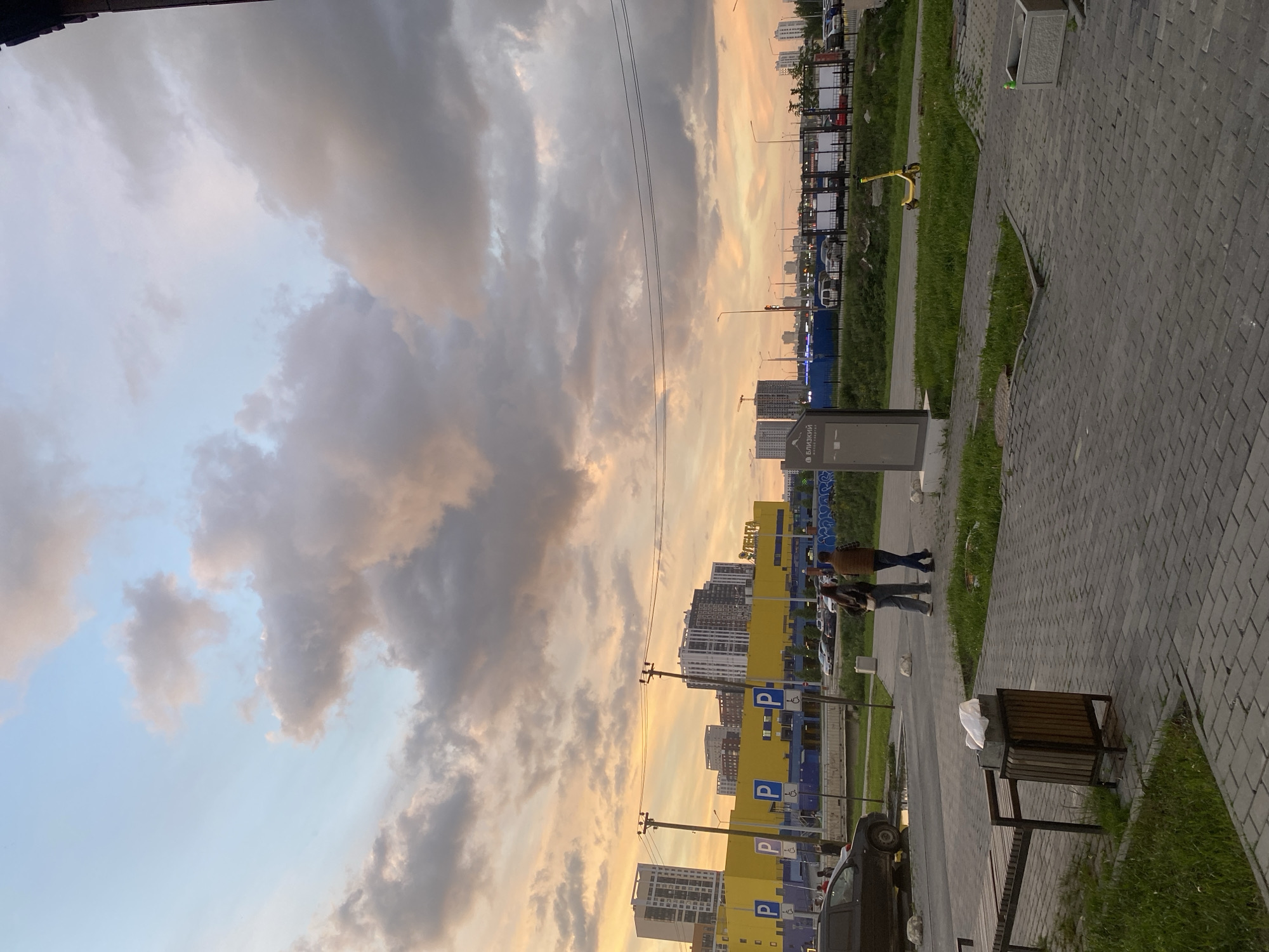 The image captures an urban street scene during sunset or golden hour, with warm light bathing the environment. The sky features dramatic clouds, displaying a blend of soft orange and pink hues from the setting sun against cooler blues and grays. Utility wires stretch across the mid-ground.
In the foreground and mid-ground, a paved pedestrian pathway made of interlocking grey bricks runs alongside grassy verges. Two individuals are walking on this path; one wears a brown jacket and blue jeans, and the other is in a darker jacket and jeans. A dark metal and wood trash receptacle, with a white bag inside, sits on the grass next to the path.
Further into the background, modern multi-story buildings, some with distinctive yellow facades, line a street or parking area. Numerous blue parking signs with a white 'P' symbol, several of which also display a white wheelchair symbol, are visible in front of these buildings. Various cars are parked in the area. A grey kiosk or information stand stands near the pathway, displaying the Russian word "БЛИЗКИЙ" (BLIZKIY). A yellow electric scooter is parked on a grassy patch further down the street. The distant cityscape includes more buildings, typical of a developed urban area. The overall scene conveys a calm, late-evening atmosphere.