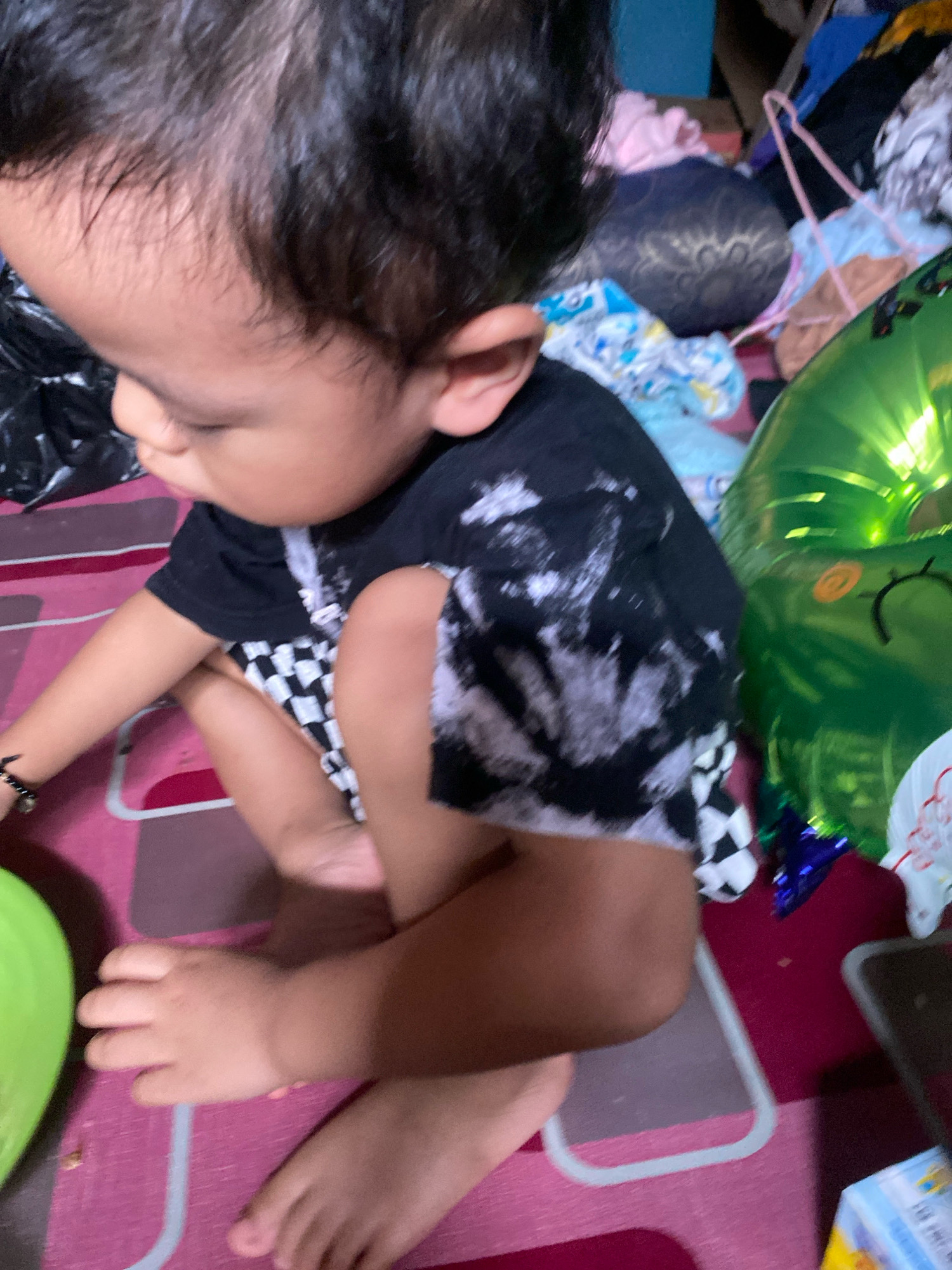 A child is sitting on a pink striped mat with a dark pattern. The child is wearing a black t-shirt with a white tie-dye pattern and black and white checkered shorts. The child's left hand is resting on their knee, and their right arm is bent at the elbow. A green balloon with a drawn-on face is visible to the right of the child. The background appears to be cluttered with various objects, including clothing and other miscellaneous items, suggesting a domestic environment. There is no specific identifiable landmark within the image to pinpoint a location within Banjarmasin, Indonesia.