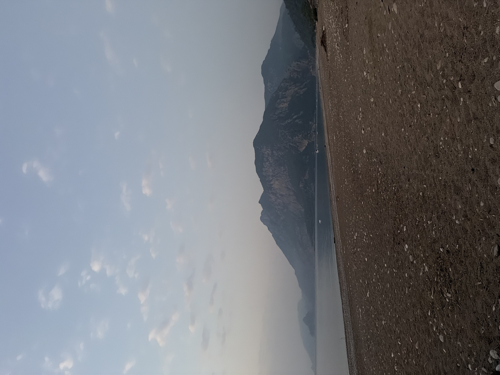 The image shows a wide, pebbly beach with dark sand leading to a calm sea. In the background, a large, rugged mountain range stretches across the horizon. The sky is a pale blue with scattered, wispy clouds. There are no people visible in the image. The scene appears to be captured during either early morning or late evening, given the soft lighting and muted colors. The environment is natural and undeveloped, suggesting a coastal area. While the specific location in Kemer, Turkey is not explicitly depicted, the mountainous coastal landscape is characteristic of the region.