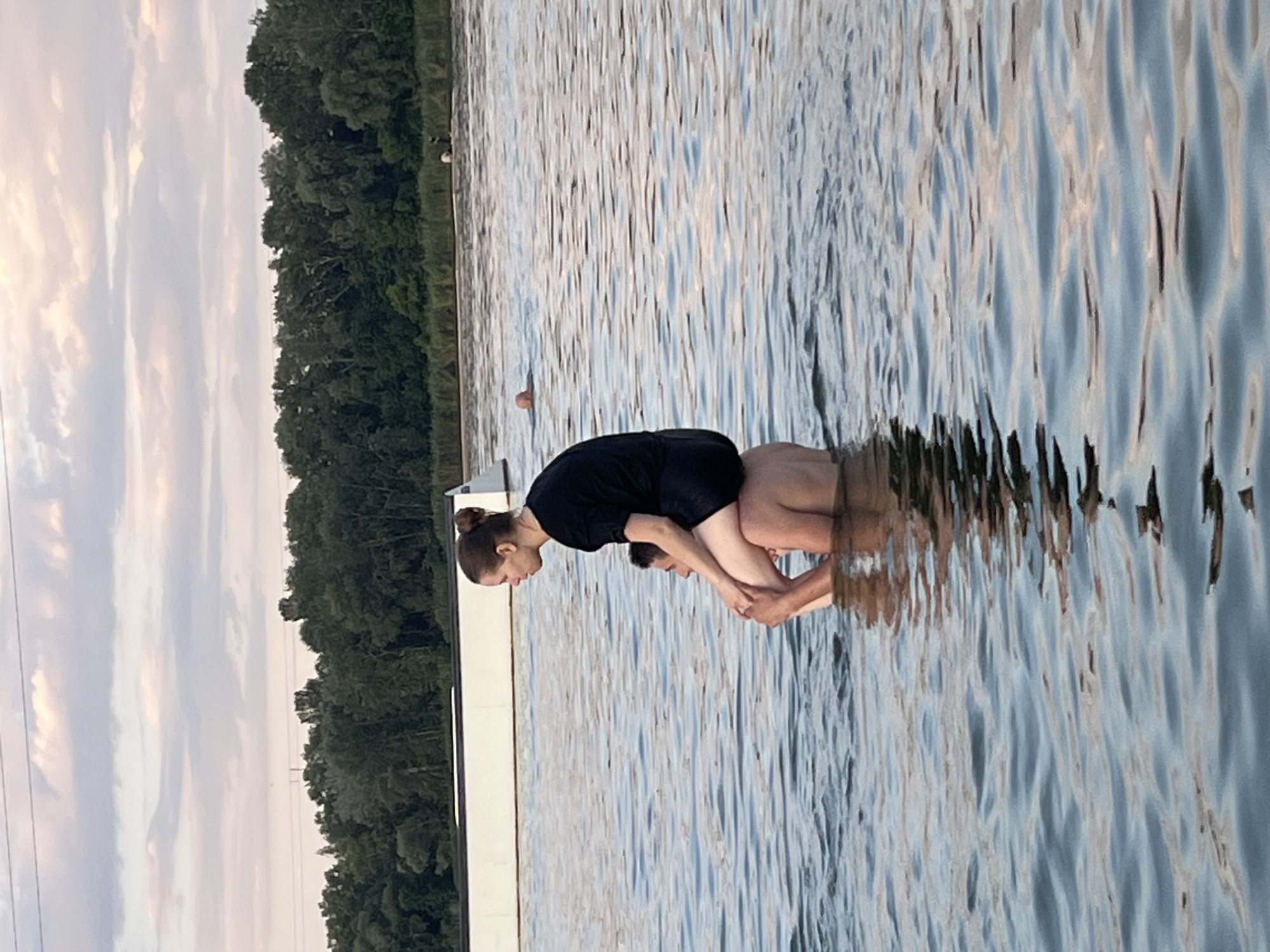 Two people are in the water near a dock. The person in the foreground is wearing a black shirt and has their hair pulled up in a bun. They are facing the other person, who is partially submerged in the water and appears to be lying on their back. The scene is outdoors with a body of water and a line of trees in the background. The sky is cloudy and appears to be late in the day, possibly sunset, due to the soft, diffused light. The water is calm with ripples reflecting the sky and surrounding trees. The overall mood is peaceful and relaxed.